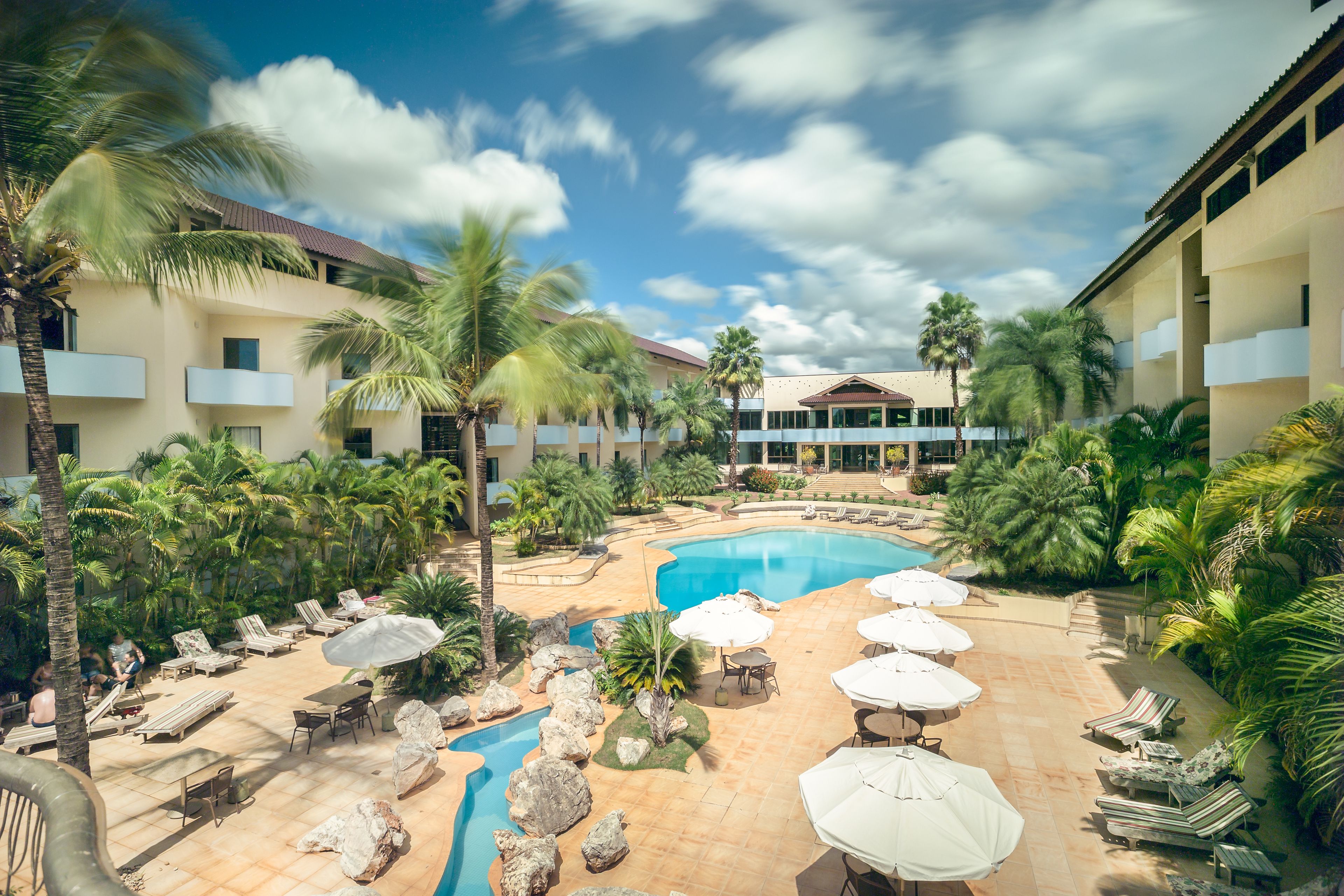 A resort pool area with lounge chairs, umbrellas, and palm trees.