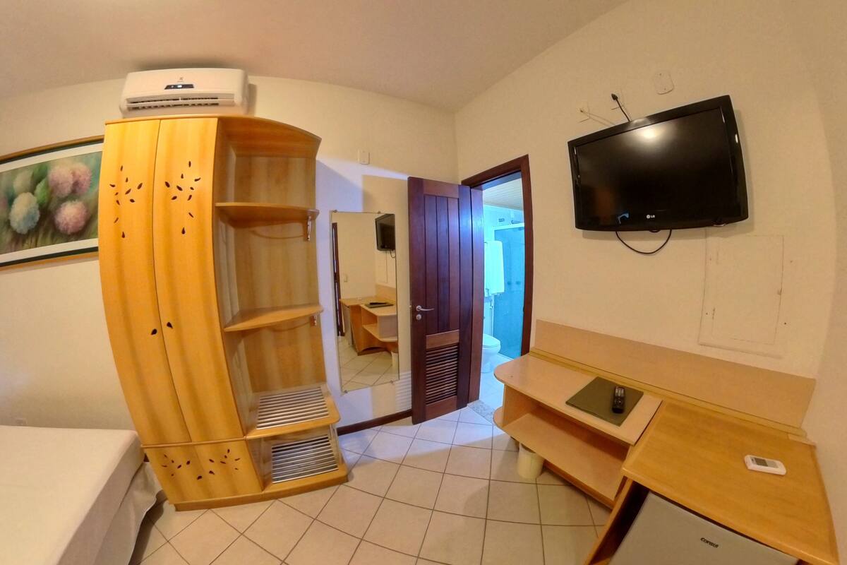 A hotel room with a bed, a wooden wardrobe, a desk, a television, and a bathroom visible through an open door.