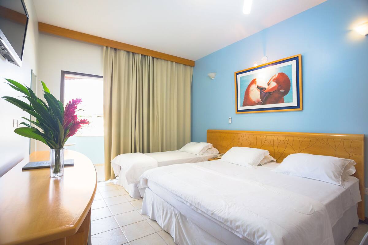 A hotel room with two beds, a wooden headboard, a painting on the wall, a desk with a plant, and a window with curtains.