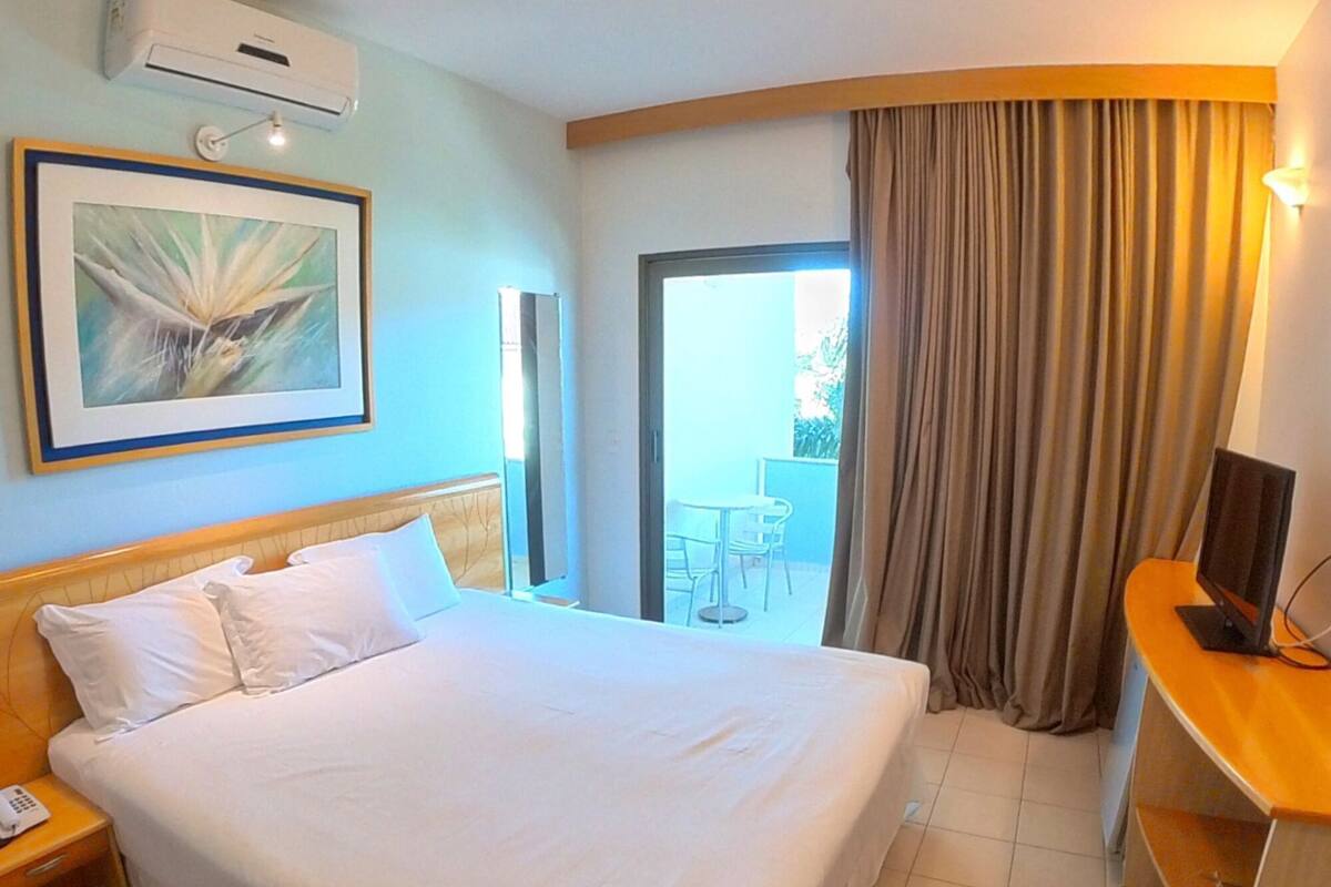 A hotel room with a bed, a desk, a TV, a painting, and a door leading to an outdoor area.