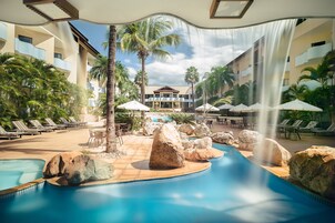 Un área de piscina con sillones de descanso, rocas grandes, palmeras y una cascada.