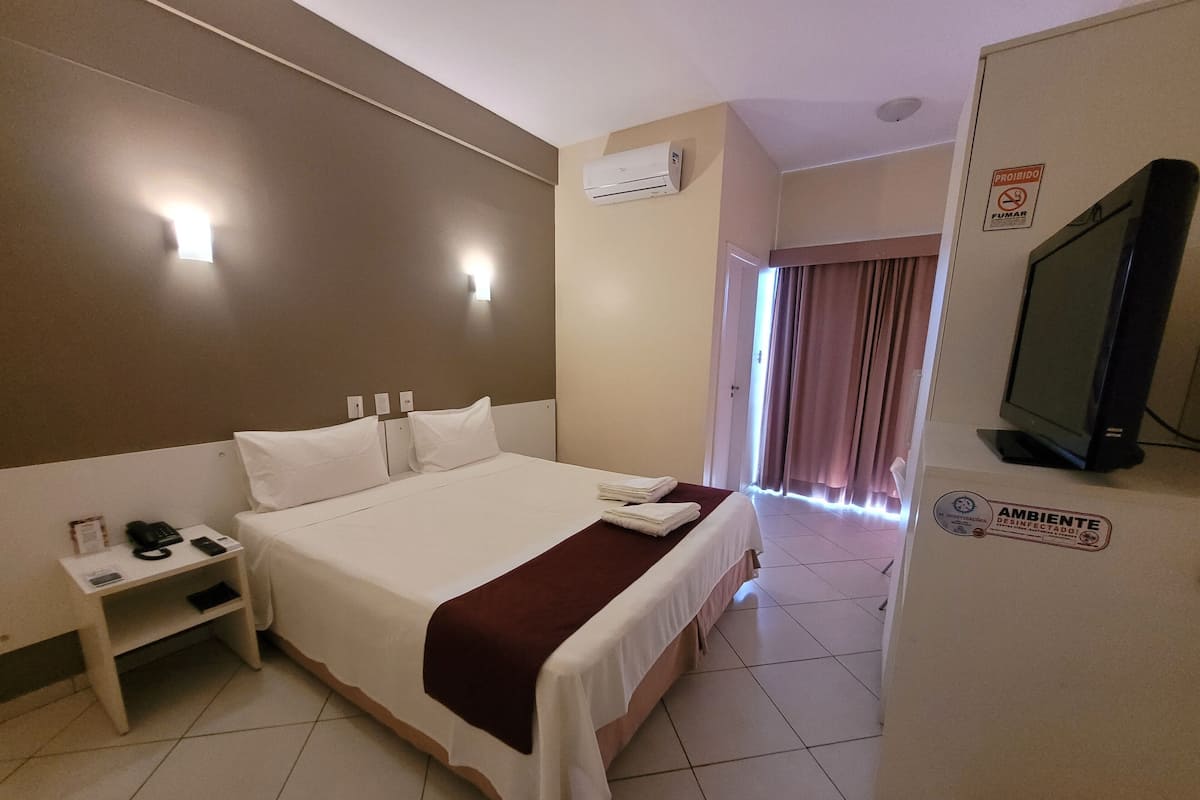 Habitación de hotel con una cama grande, una mesita de noche con un teléfono y una lámpara, un equipo de aire acondicionado, un televisor y un letrero que indica que la habitación está desinfectada.