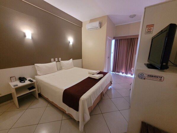 Une chambre d’hôtel avec un grand lit, une table de chevet avec un téléphone et une lampe, un appareil de climatisation, un téléviseur et un panneau indiquant que la chambre a été désinfectée.