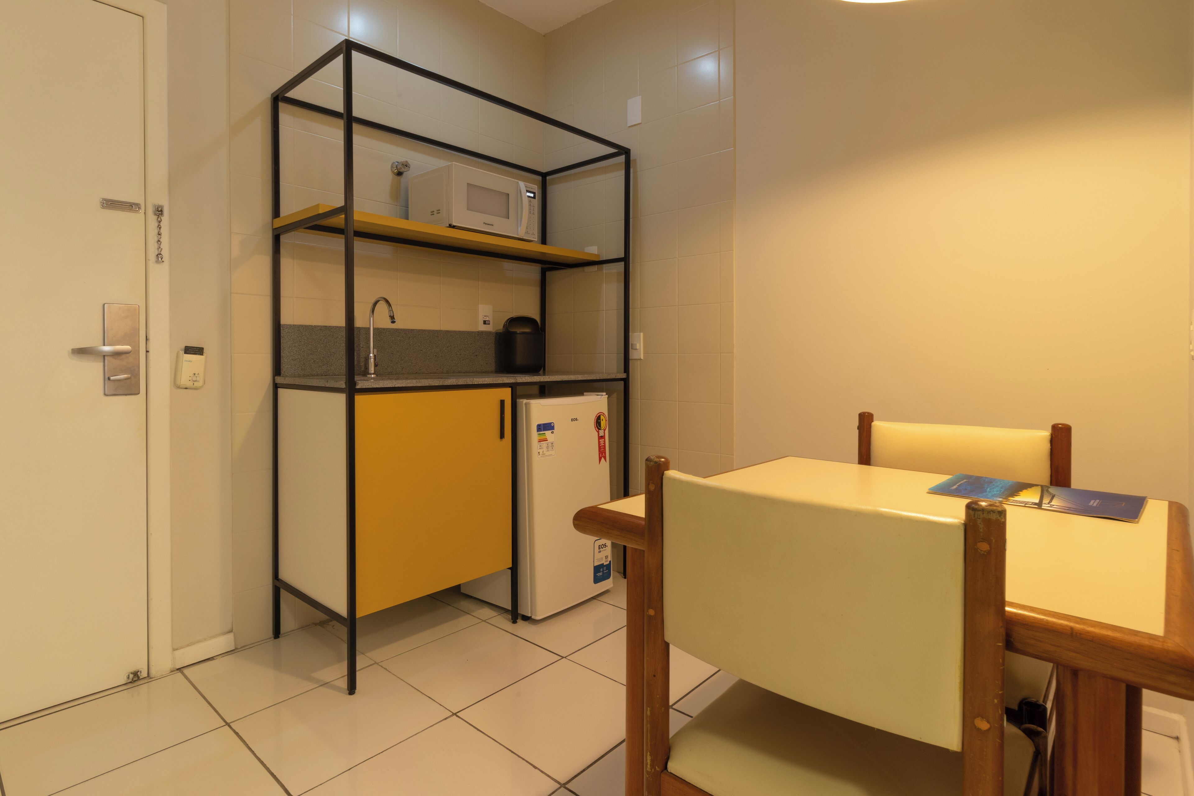 Cocina compacta con microondas, lavamanos y refrigerador. Hay una silla de madera y una mesa pequeña con un libro.