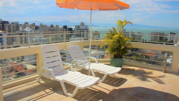 A balcony with two white lounge chairs, an orange umbrella, and a potted plant. - Rio Branco Apart Hotel (Florianópolis)