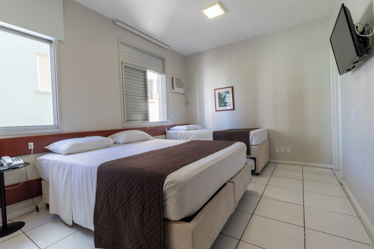 A hotel room with a large bed, a smaller bed, a bedside table with a phone, a TV mounted on the wall, and a window with blinds.