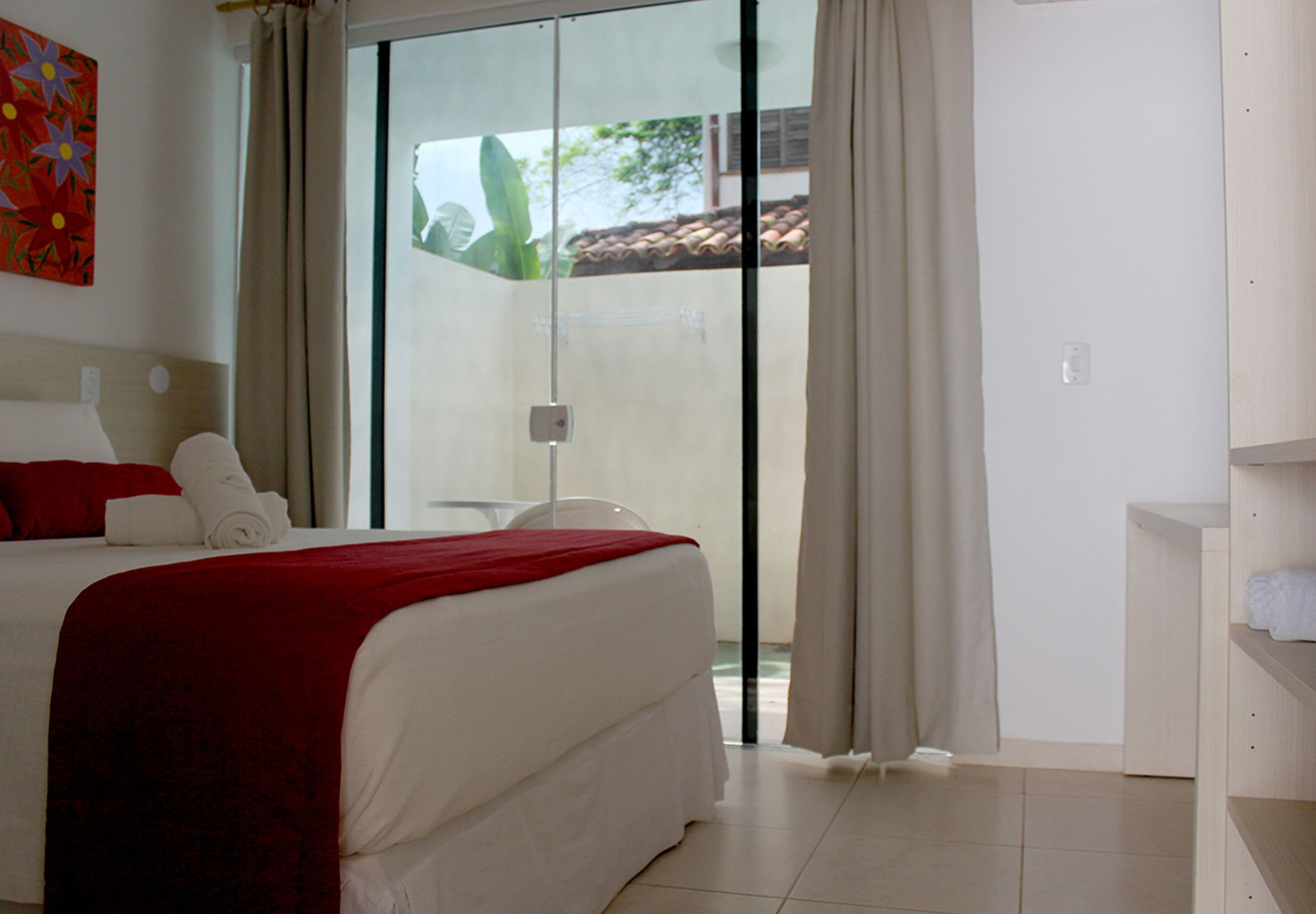 A hotel room with a bed, a red blanket, a white pillow, a glass door leading to a balcony, and a wooden shelf with a towel.