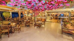 A spacious indoor area with multiple wooden tables and chairs, a central bench with cushions, and a ceiling adorned with artificial flowers.