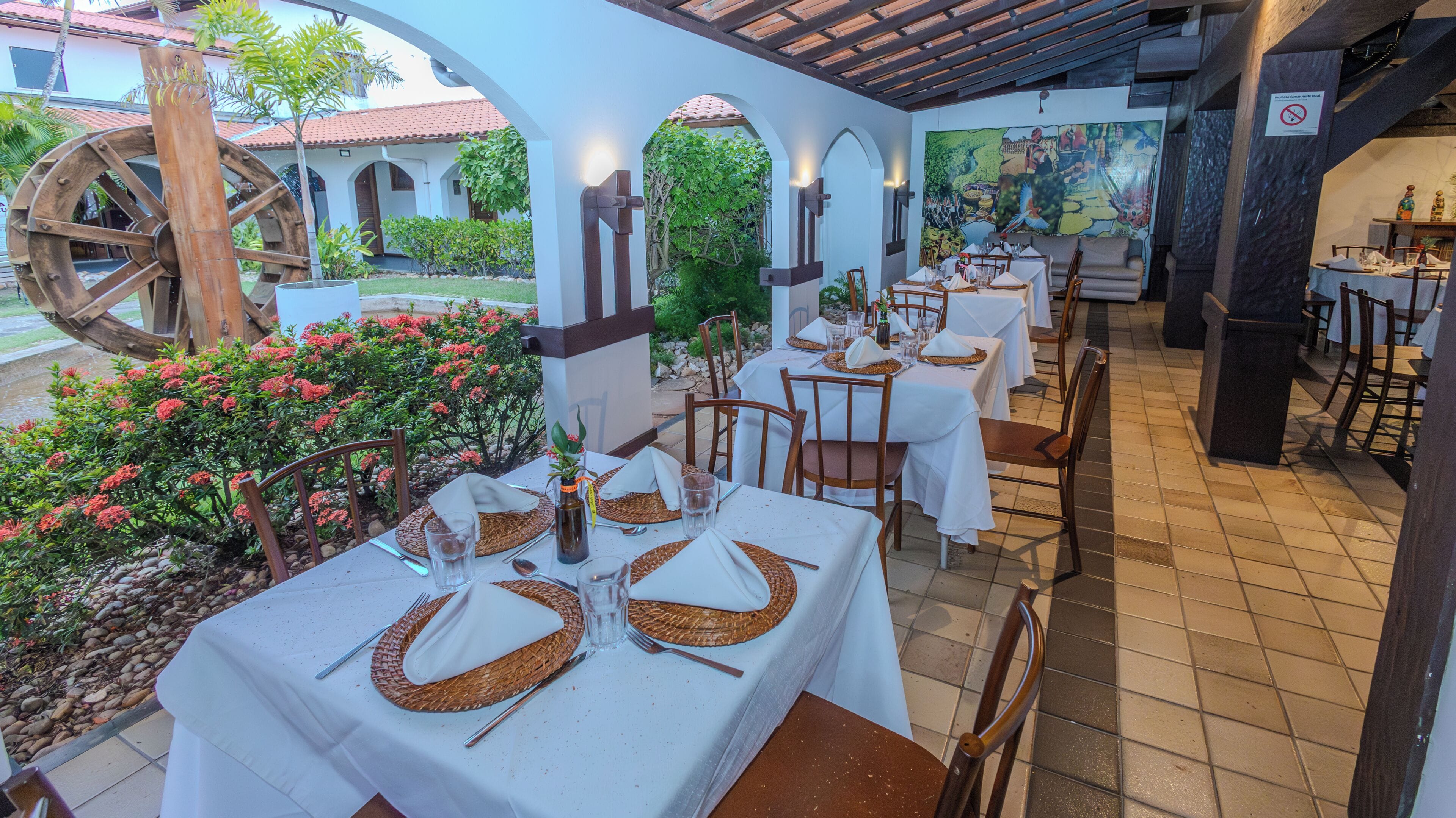 Un área de comedor al aire libre con mesas dispuestas para dos, rodeada de arcos y un mural.