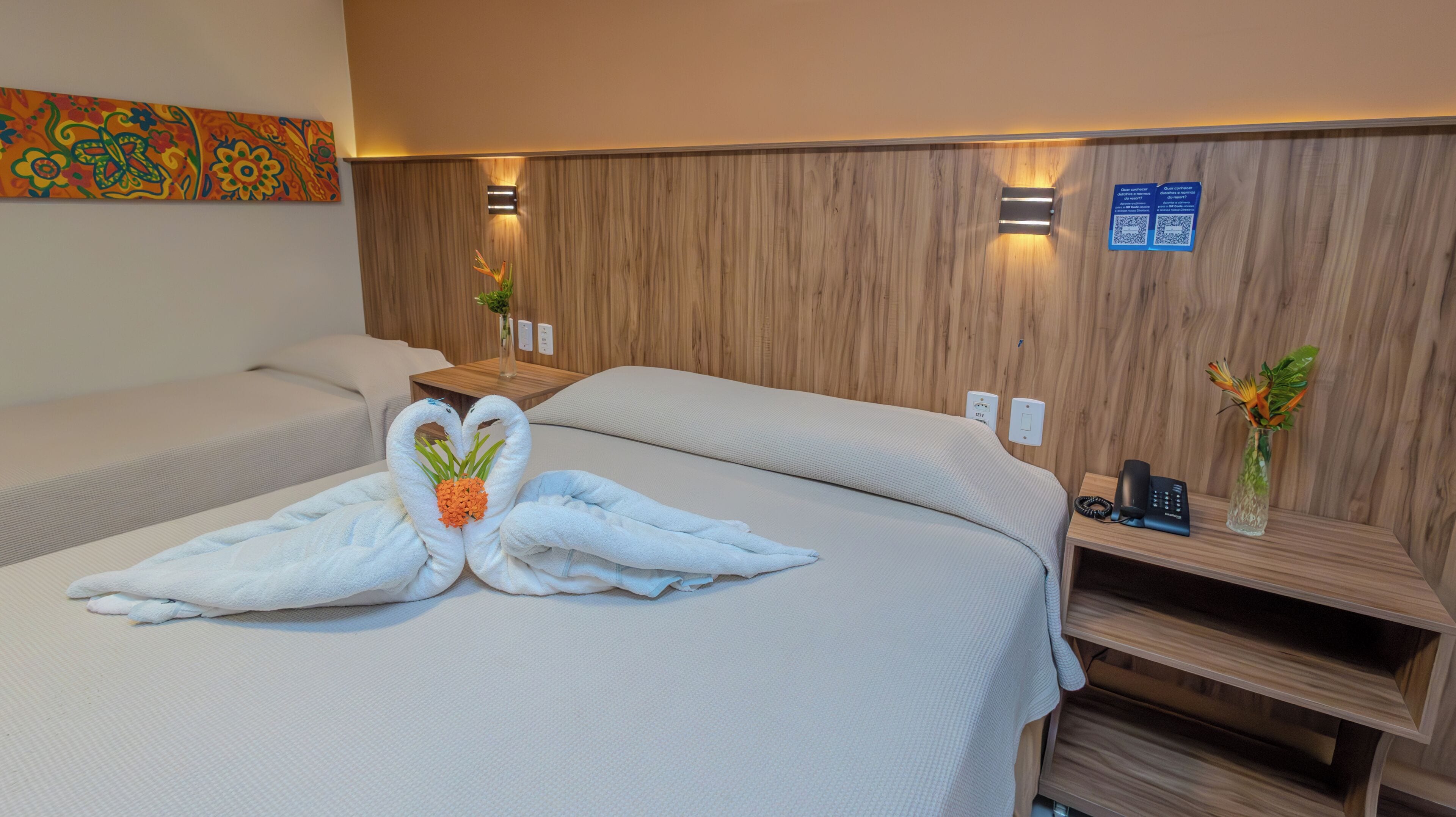 A hotel room with two beds, a wooden wall panel, a small table with a vase, and a phone on a wooden shelf.