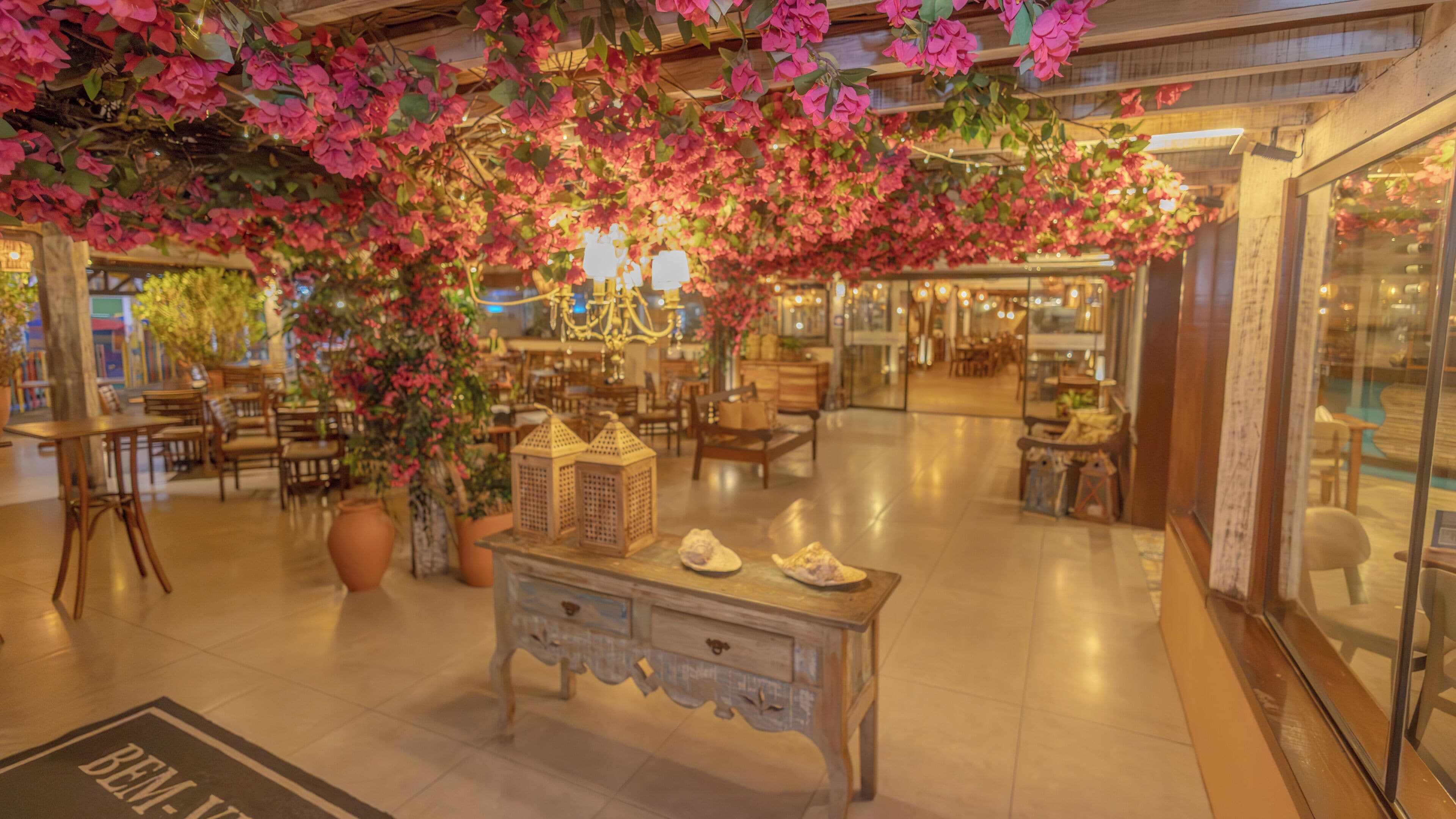 Un vestíbulo amplio con una zona decorativa central que presenta un gran macetero, una mesa de estilo vintage con zapatos y flores rosas colgantes.