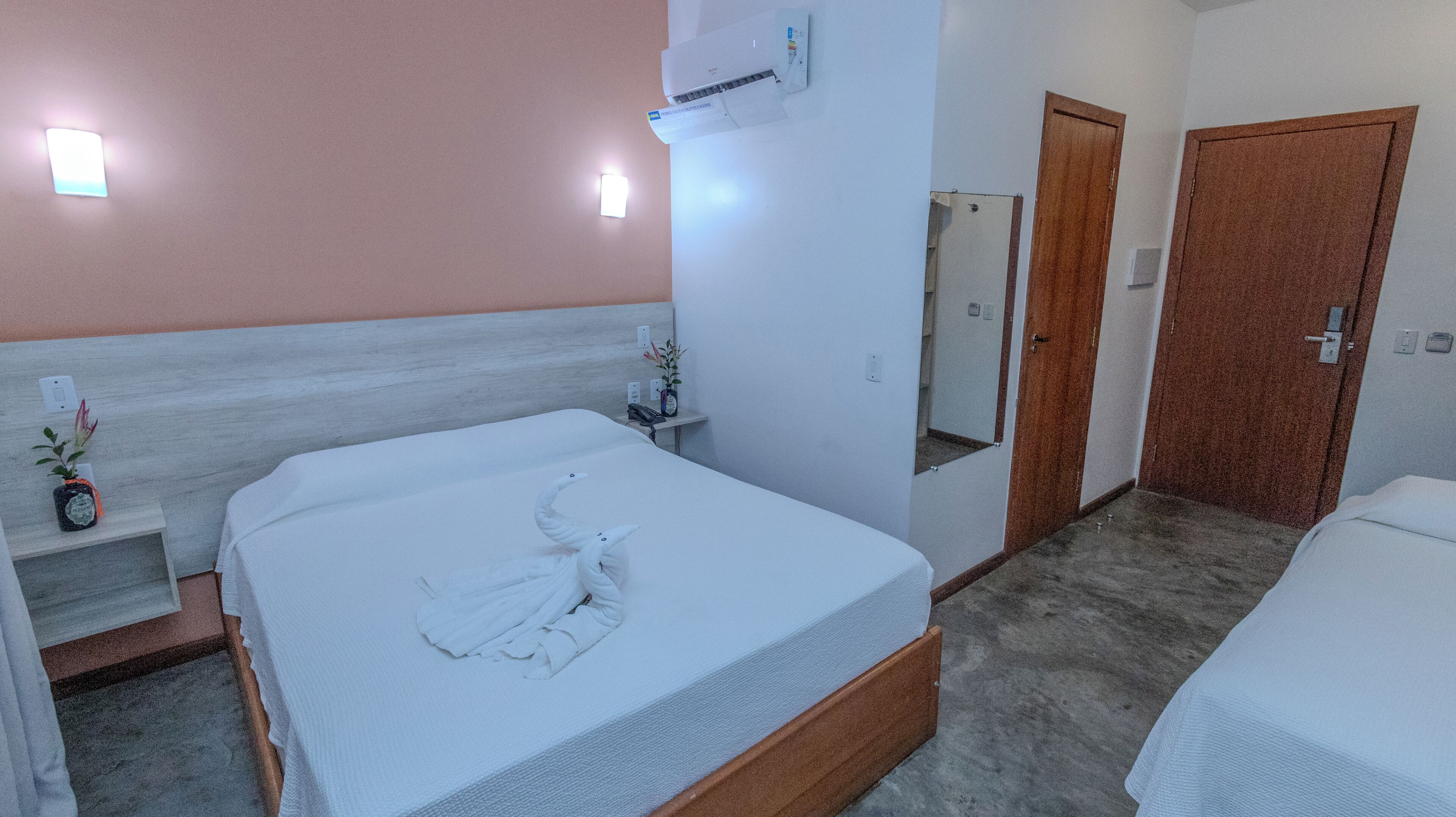 A hotel room with a bed, a bedside table with a phone, a wall-mounted air conditioner, a mirror, and a door.