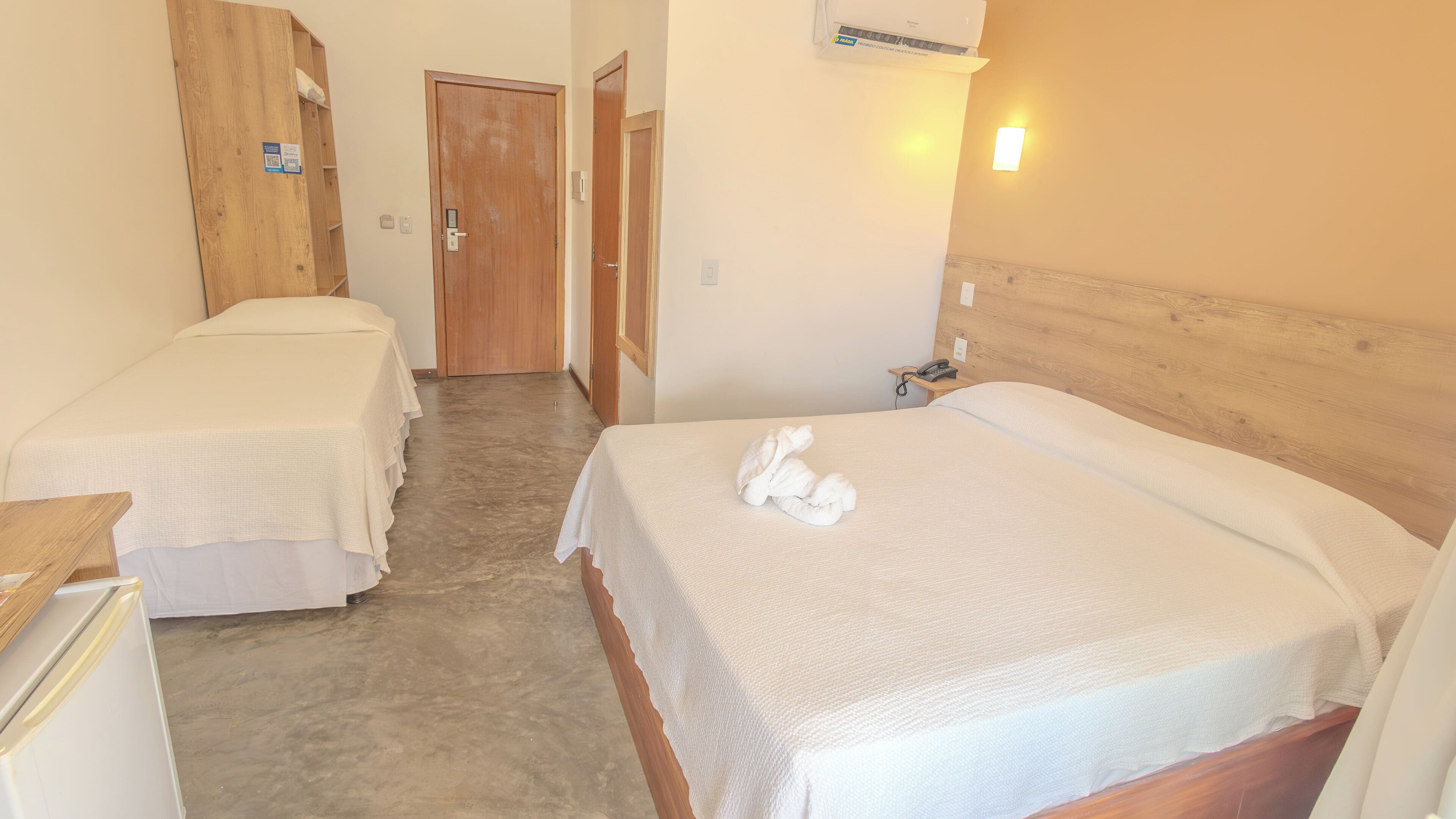 A hotel room with two single beds, a wooden wardrobe, a small desk with a telephone, and a wall-mounted air conditioner.