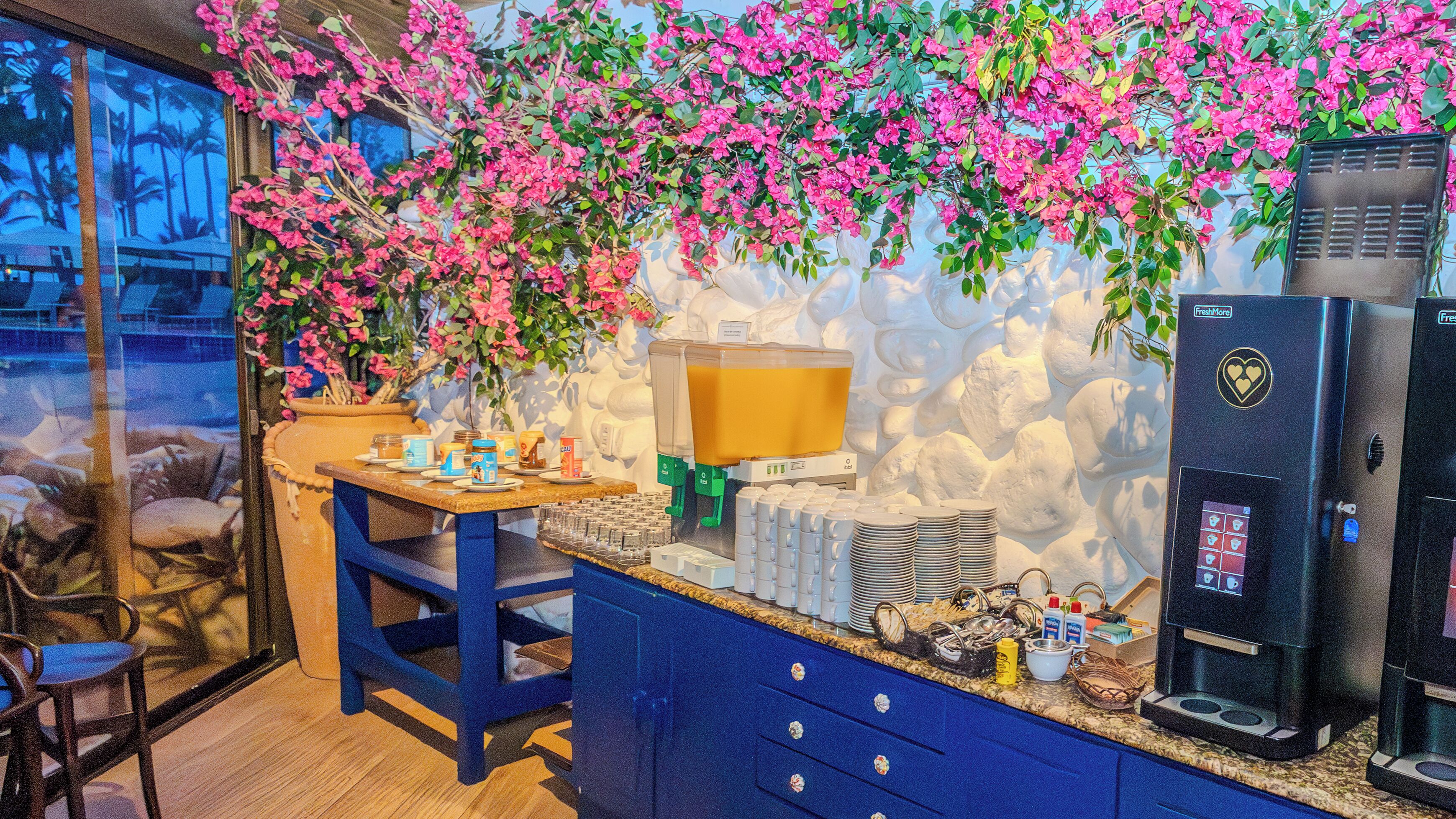 Un área de cocina con una cafetera, un dispensador de zumo de fruta y una mesa con varios objetos.