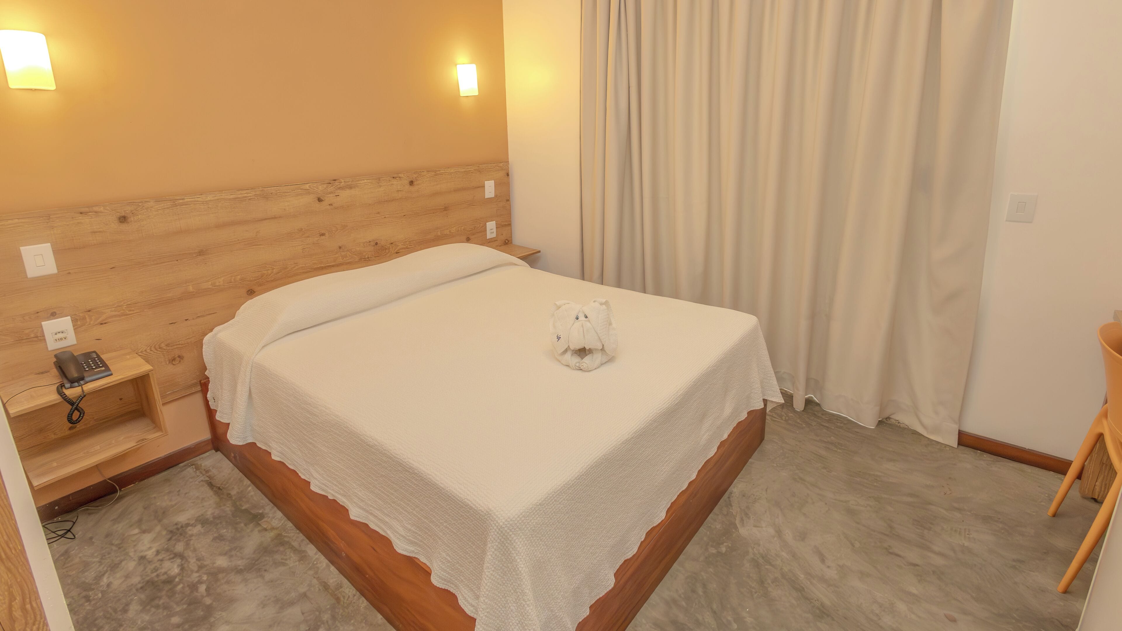 A neatly made bed with a white bedspread and a towel shaped like an elephant. There is a wooden nightstand with a phone and a power outlet.
