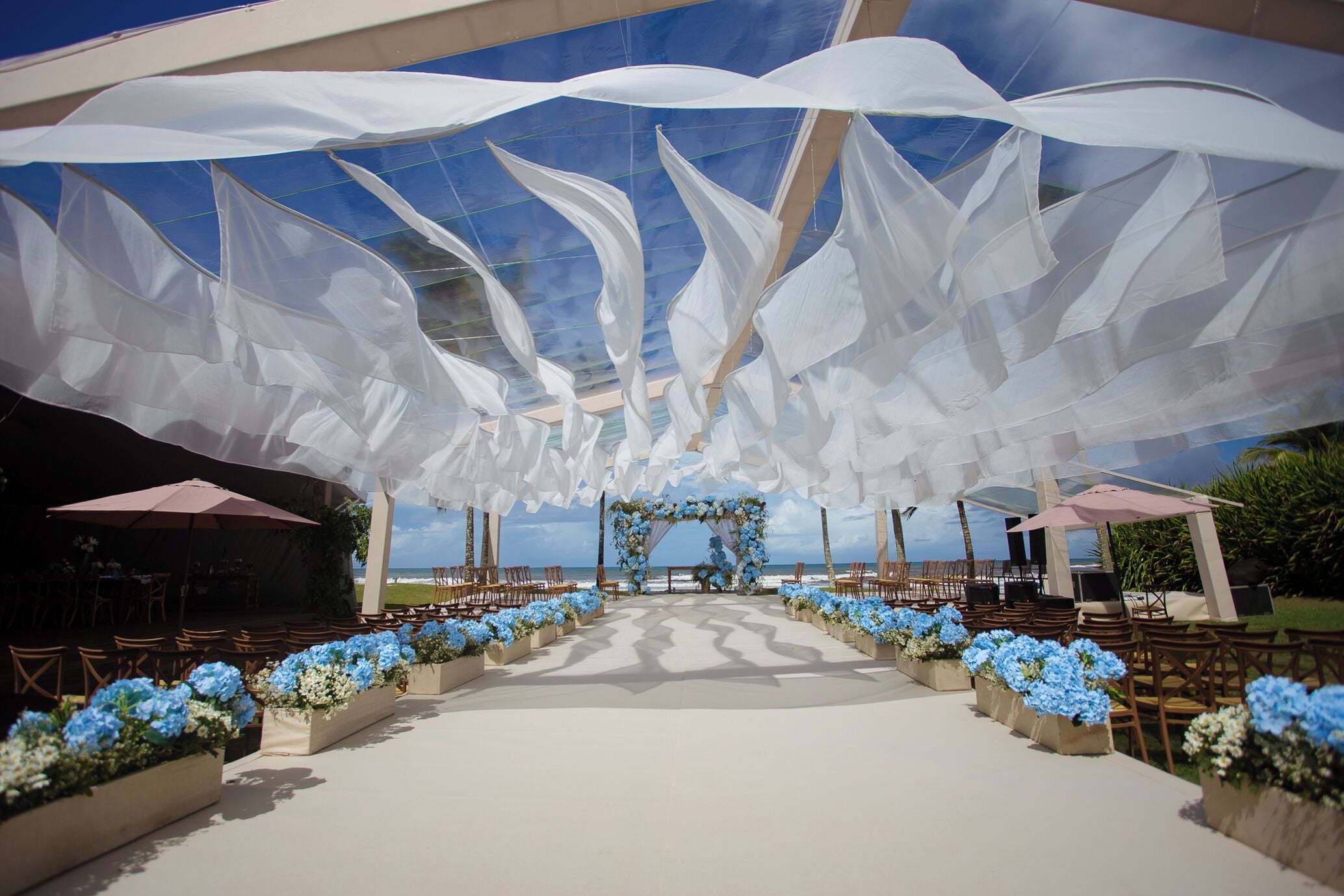Un salón de bodas al aire libre con pasillo blanco, arreglos florales y vistas al océano.