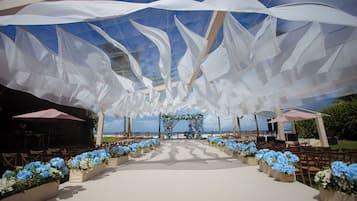 Un salón de bodas al aire libre con pasillo blanco, arreglos florales y vistas al océano.