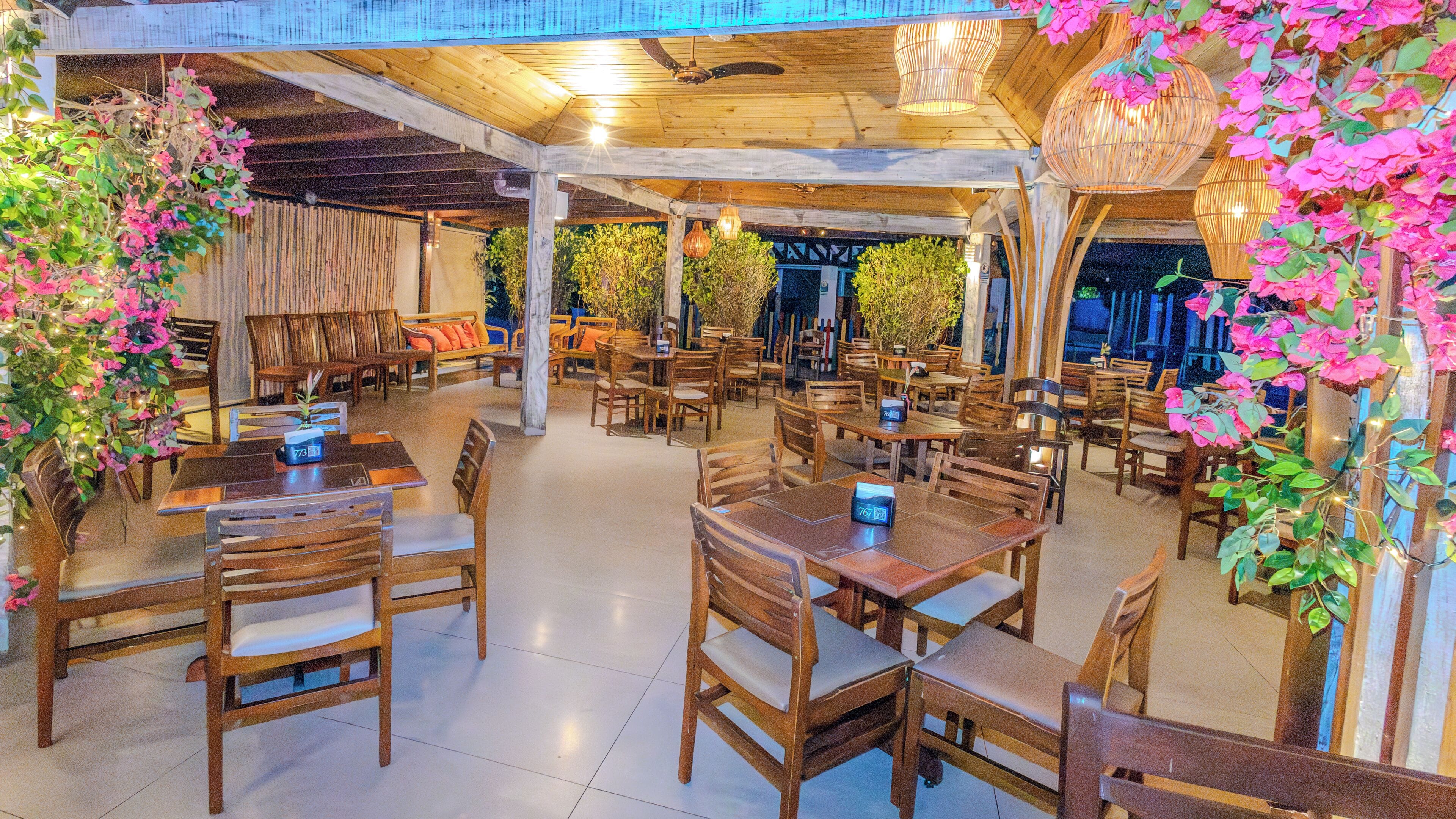 A spacious indoor dining area with wooden tables and chairs, a ceiling fan, and hanging plants.