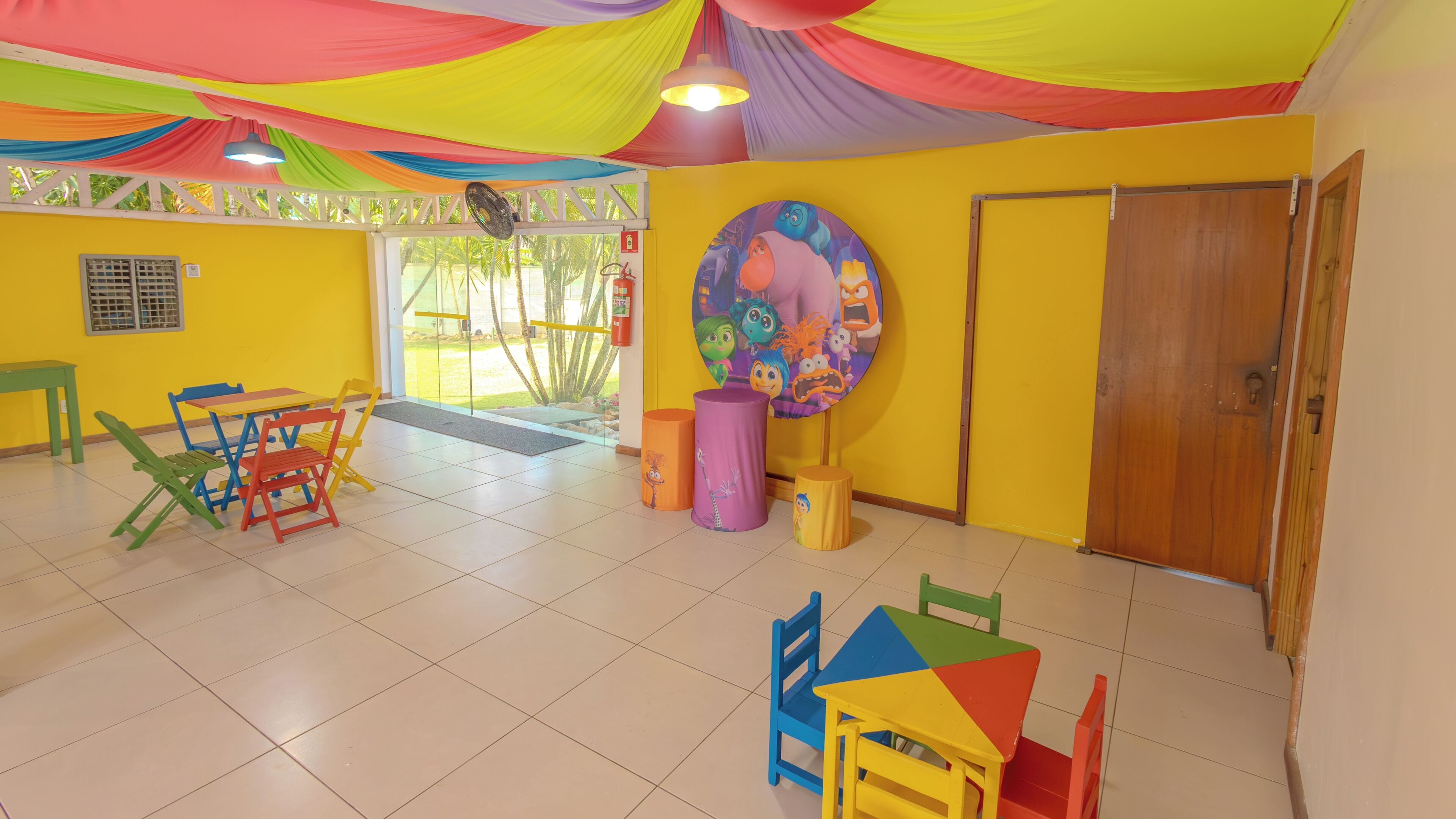 A brightly colored room with a yellow wall, a wooden door, and a colorful mural. There are several chairs and tables arranged for seating.