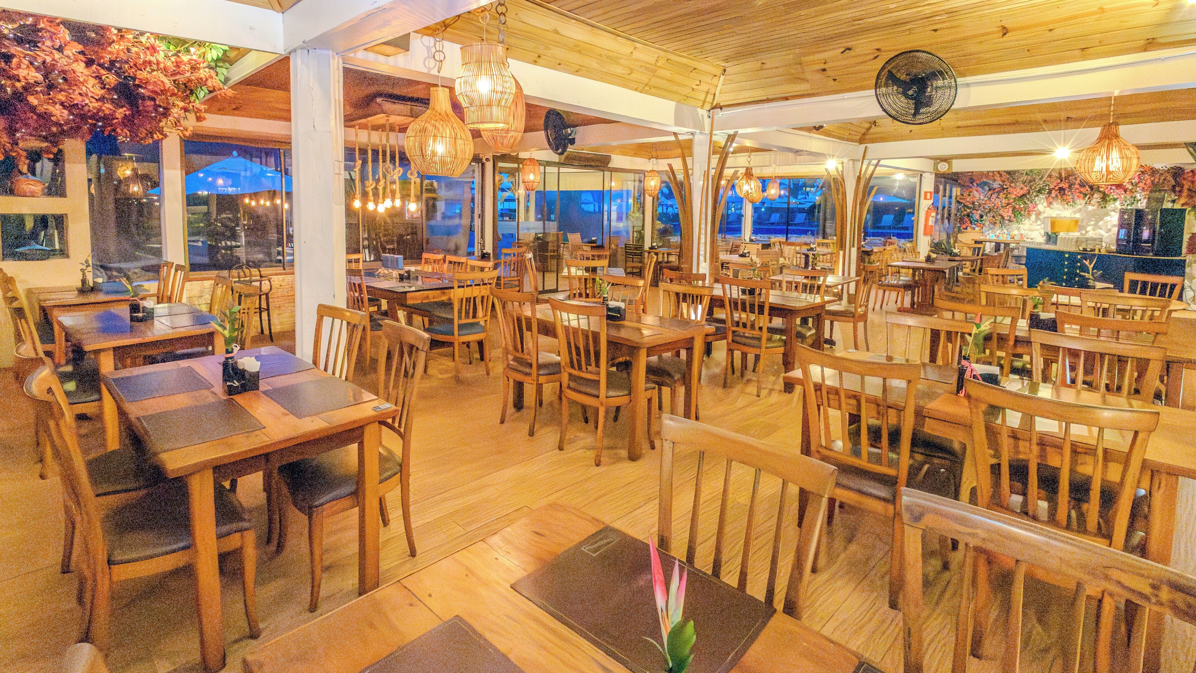 Un restaurante con mesas y sillas de madera, luces colgantes y amplios ventanales.