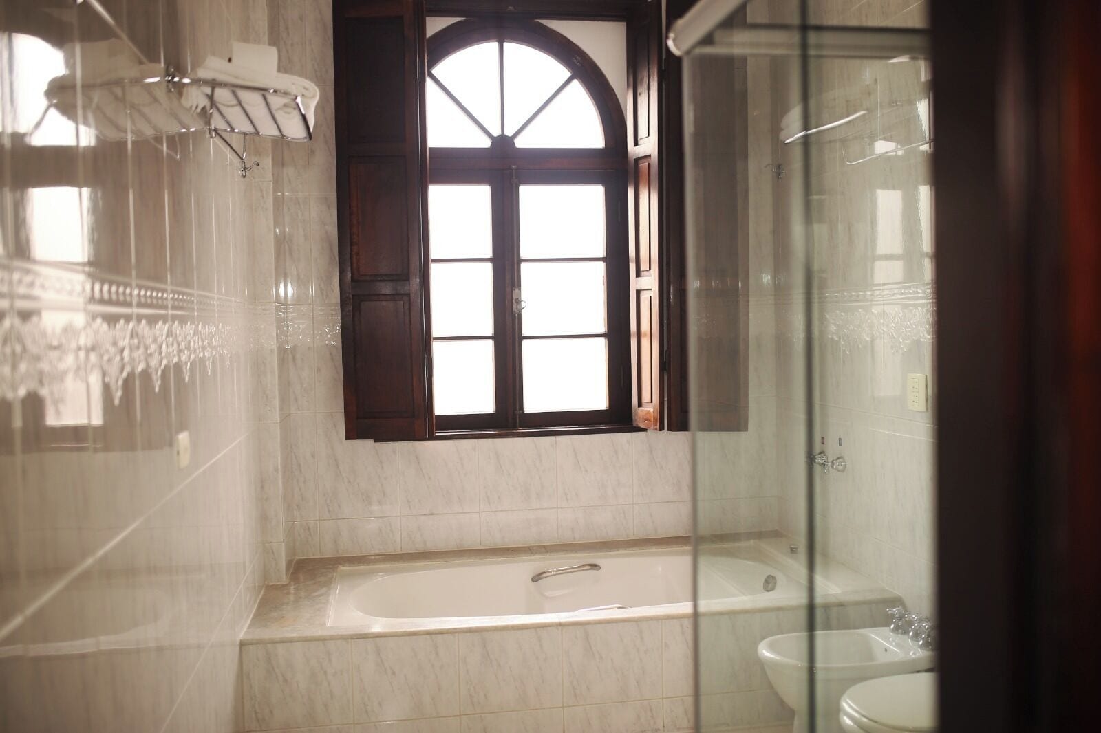 Un baño con bañera, una ventana con contraventanas de madera y mampara de vidrio.