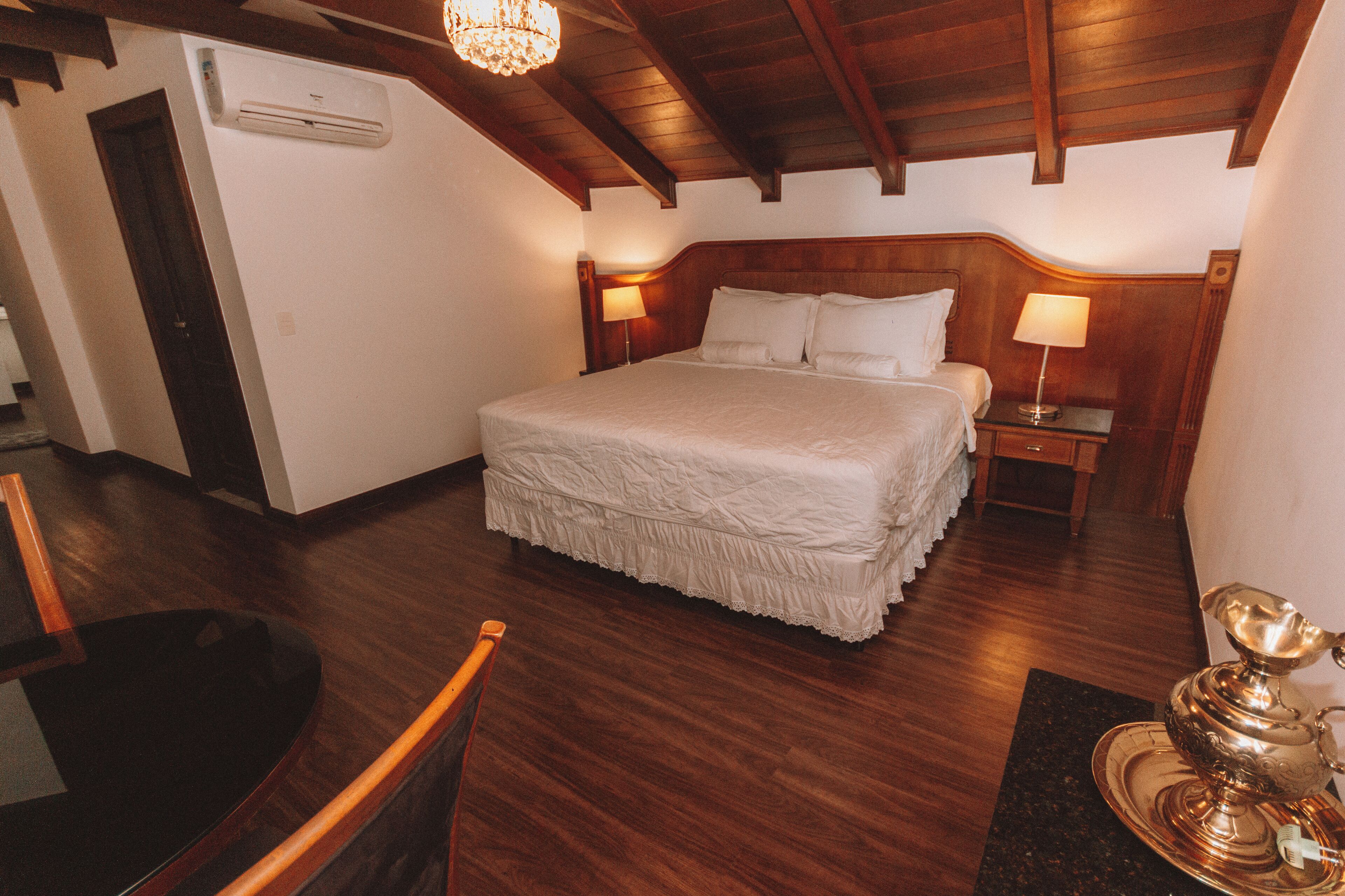 Un dormitorio con techo de madera, una cama con ropa de cama blanca, una mesita de noche con lámpara y una silla de madera.