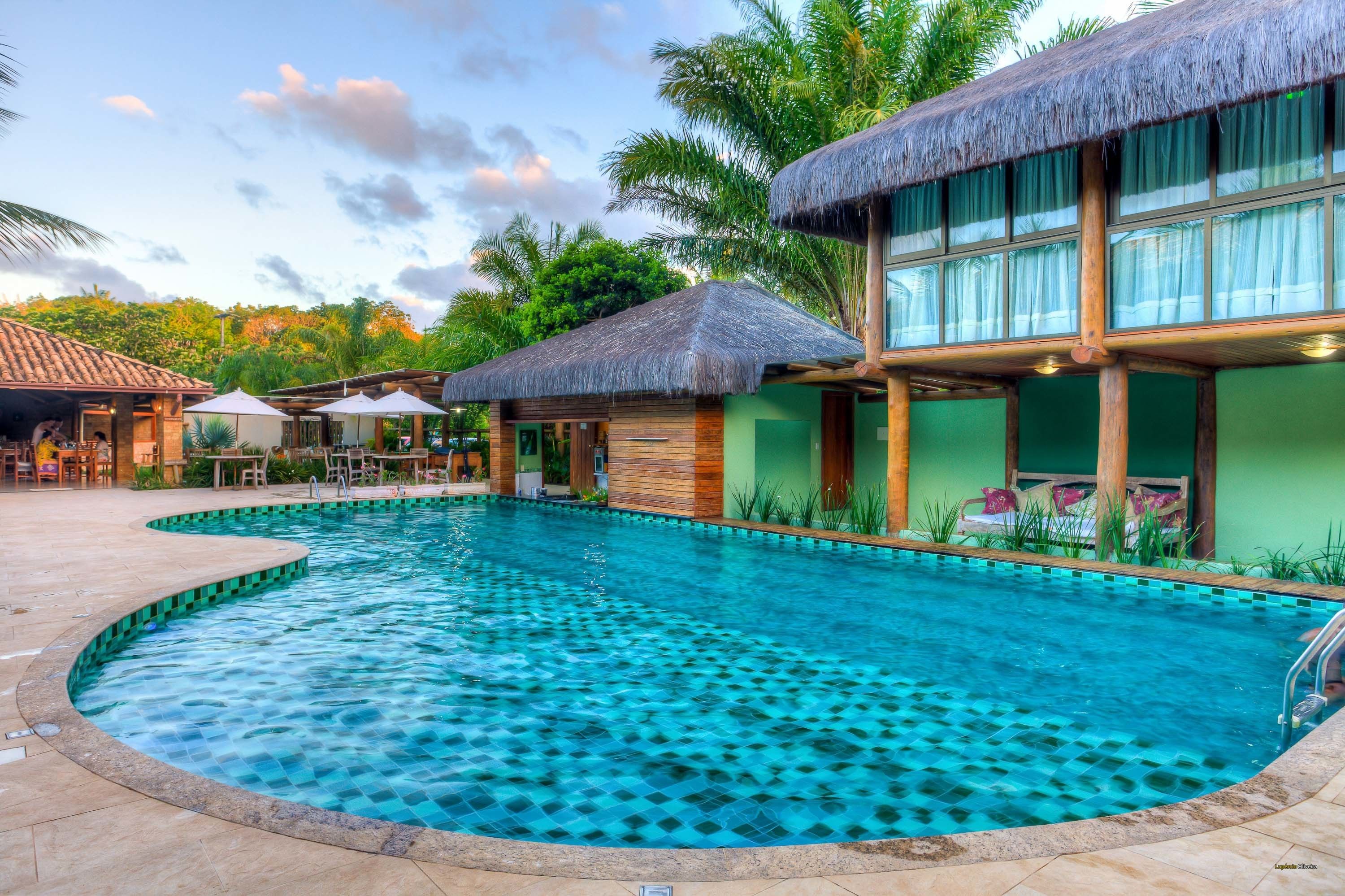 A swimming pool with a tiled edge, surrounded by a lounge area with tables and chairs, and a building with large windows and a thatched roof.