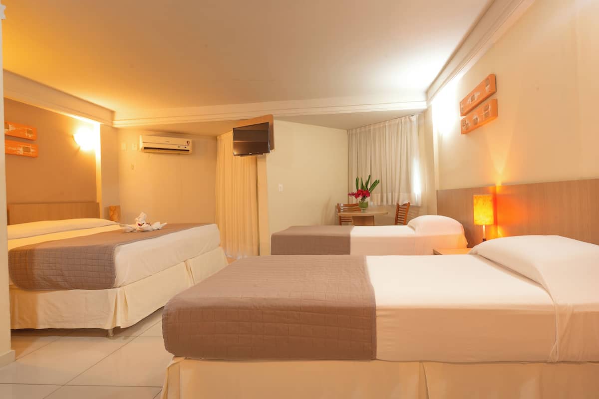 A hotel room with three beds, a flat-screen TV, an air conditioner, a small table with a vase of flowers, and a lamp.