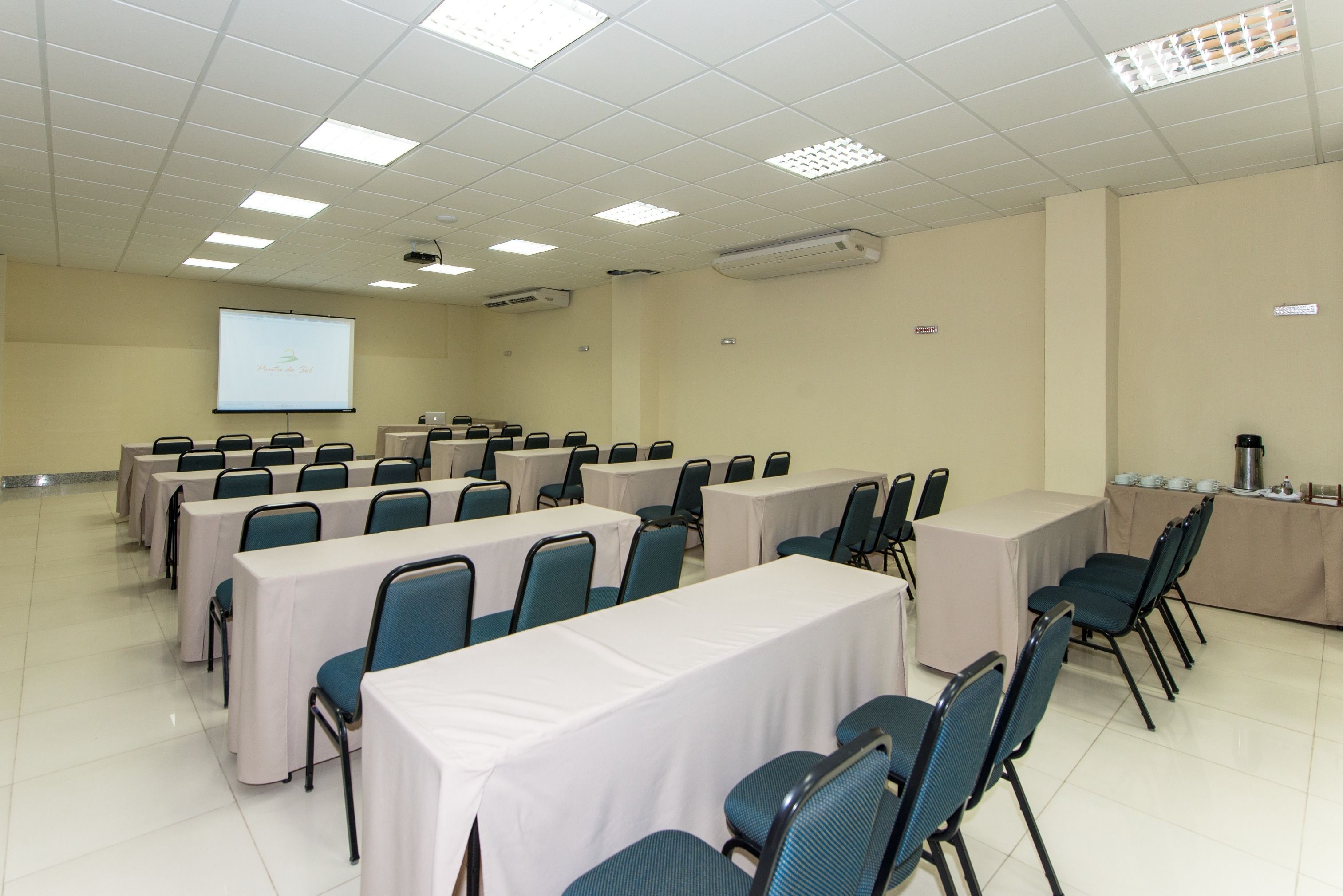 A conference room with multiple tables and chairs, a projector screen, and an air conditioning unit on the ceiling.