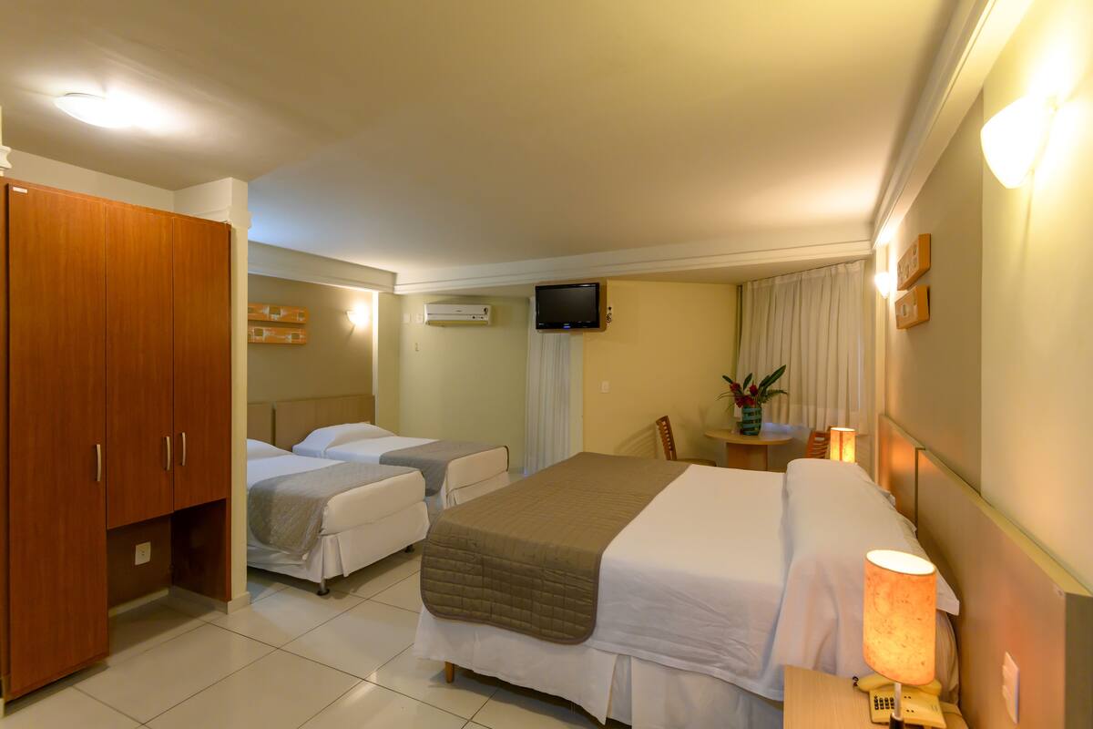 A hotel room with two beds, a wooden wardrobe, a TV, and a bedside table with a lamp.