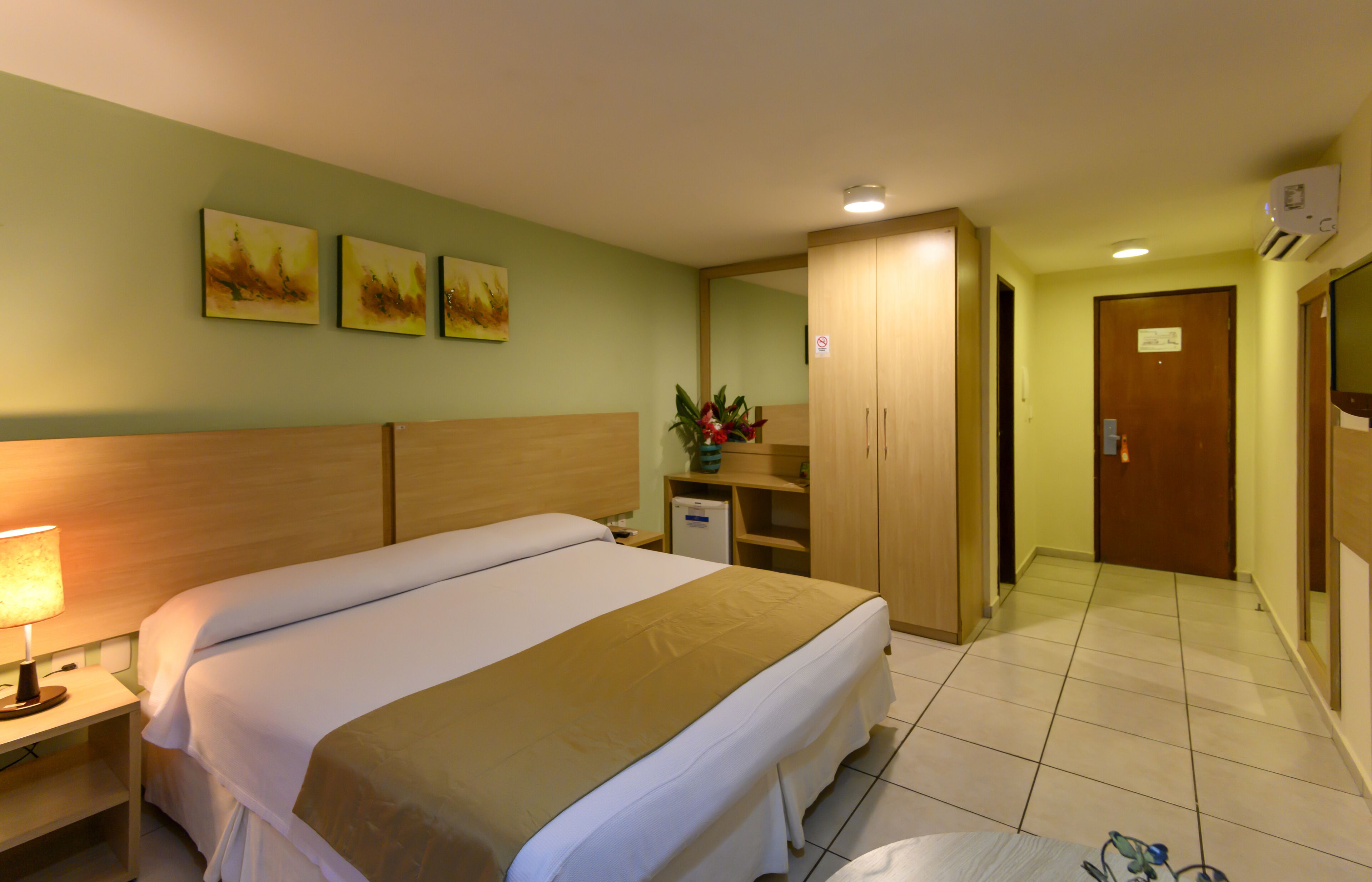 A hotel room with a large bed, a nightstand, a lamp, a small desk, a mirror, a wardrobe, and a door.