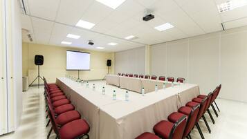 Un salón de conferencias con una larga mesa, sillas rojas, una pantalla de proyector y un orador.