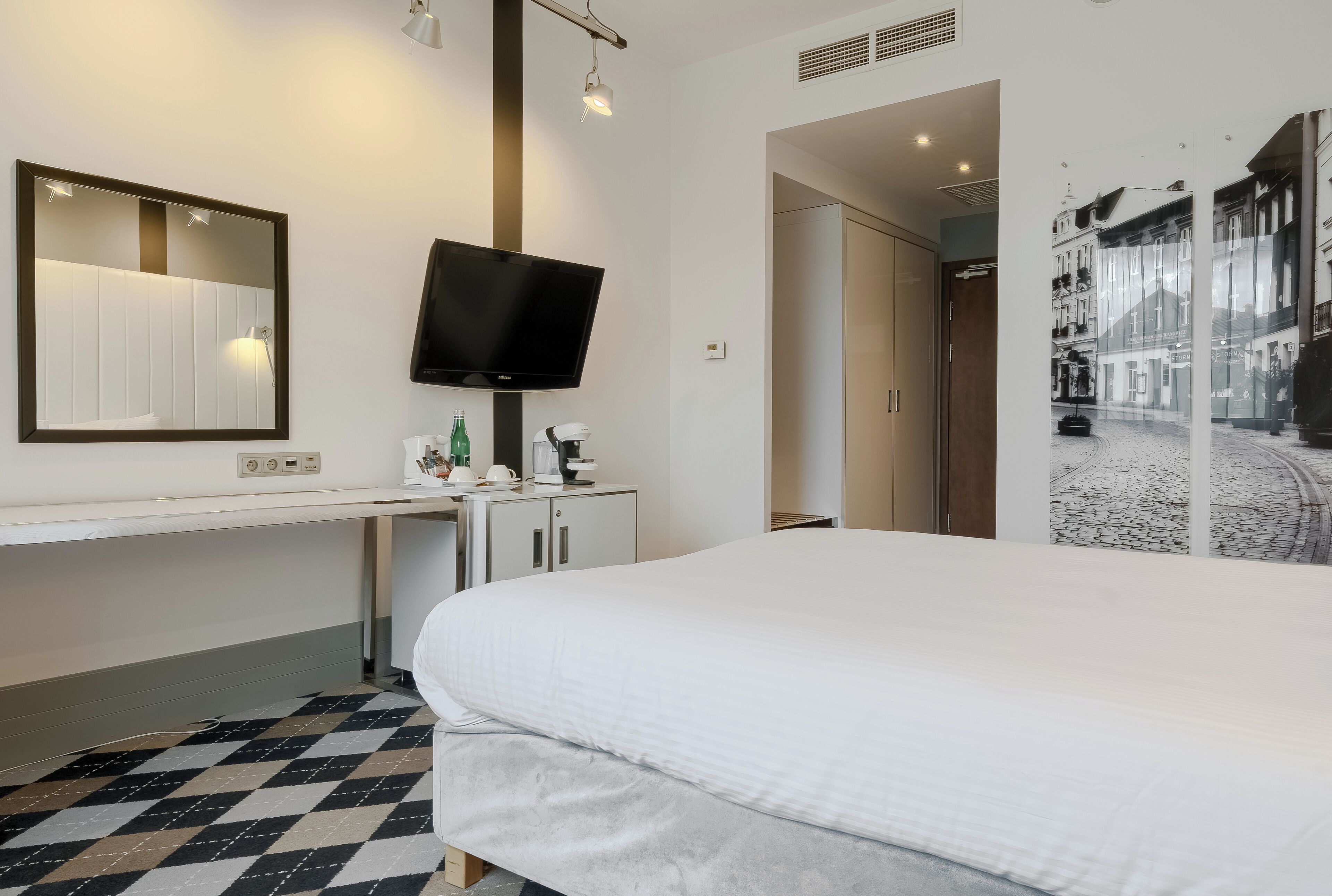 A hotel room with a bed, a desk with a flat-screen TV, a mirror, and a wall-mounted shelf with toiletries.