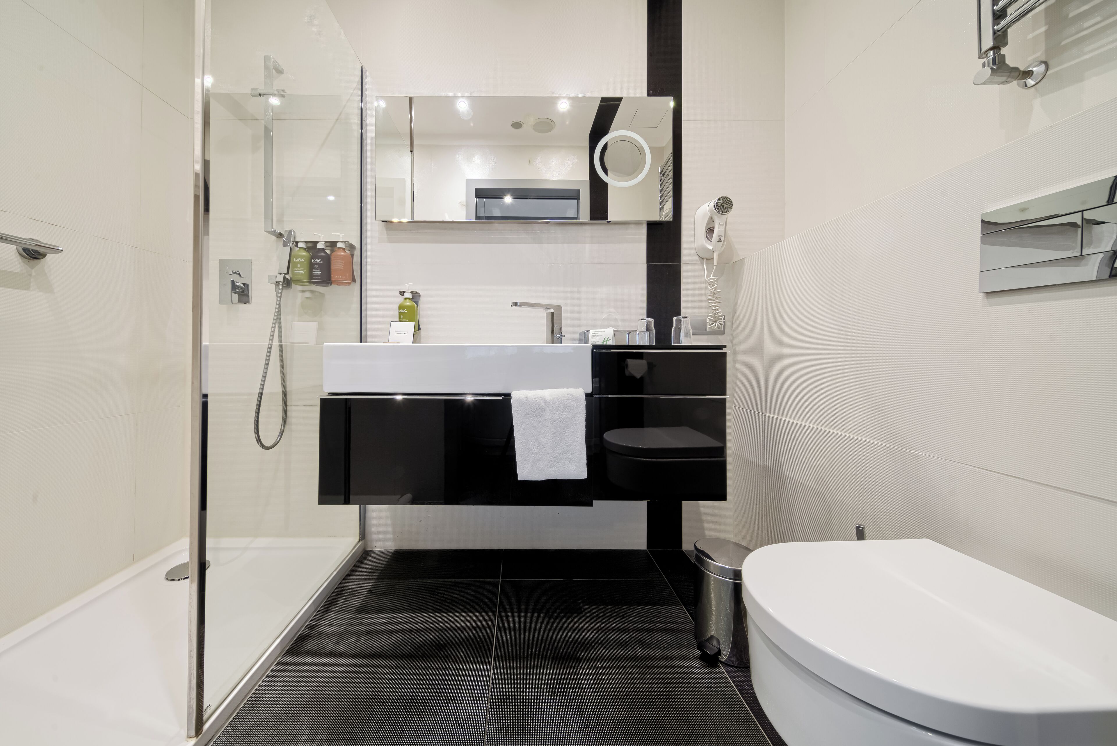 Un baño moderno con cabina de ducha de vidrio, lavamanos blanco, tocador negro, inodoro y un cubo de basura.