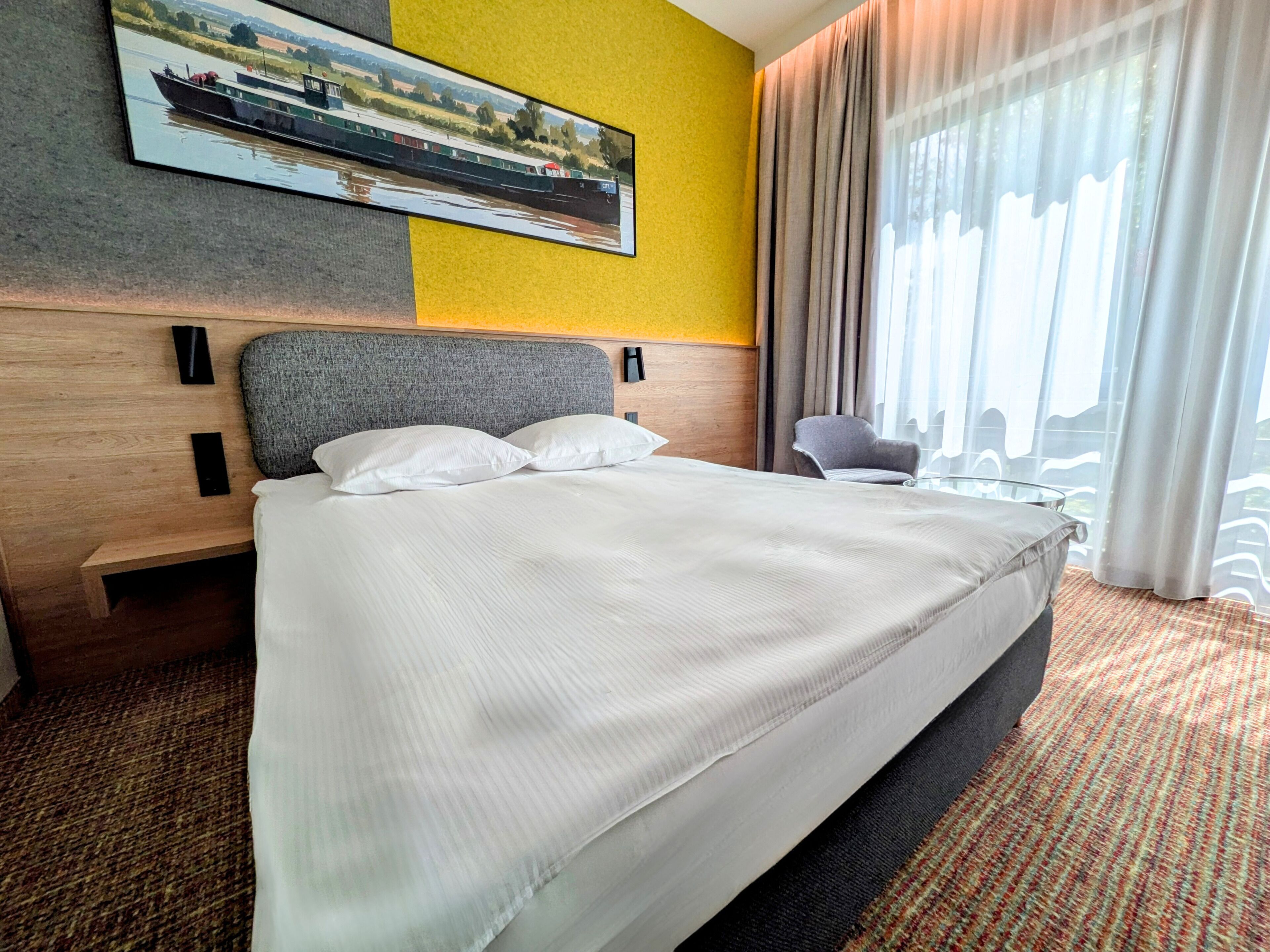 A hotel room with a large bed, a headboard, a painting of a boat, a chair, and a window with curtains.