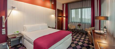Une chambre d’hôtel avec un grand lit, un bureau avec une chaise, une petite table avec un vase et un grand miroir.