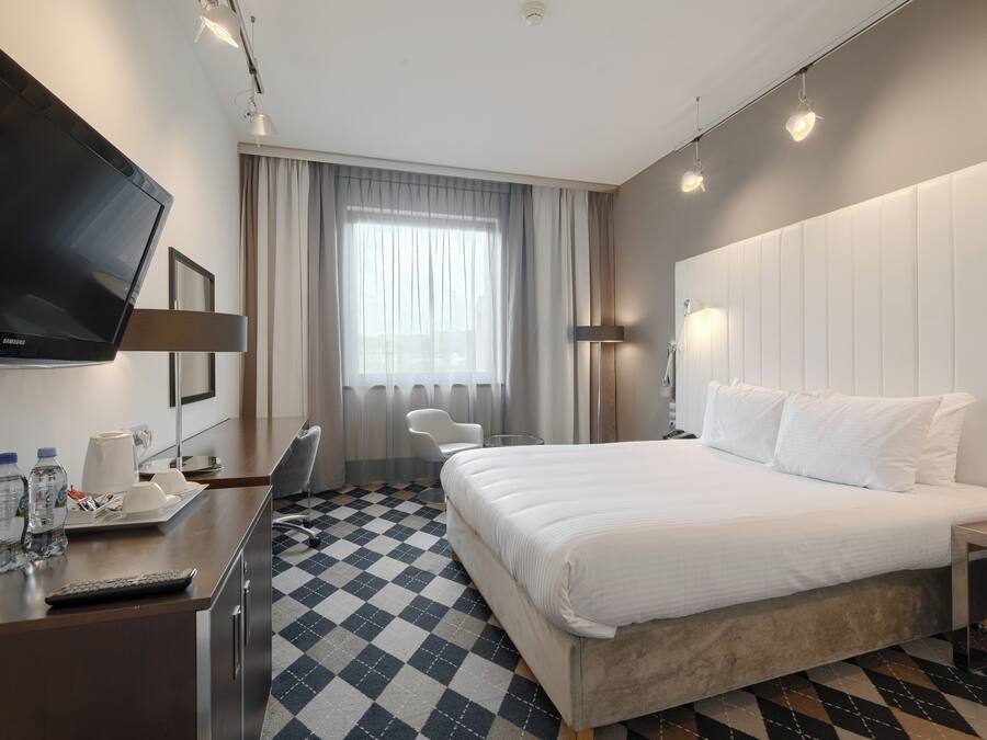 A hotel room with a large bed, a desk with a TV, a chair, a lamp, and a check-in counter.