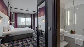 Una habitación de hotel moderna con suelo a cuadros, una cama grande, un escritorio, una silla, un espejo, una ducha y un lavabo.