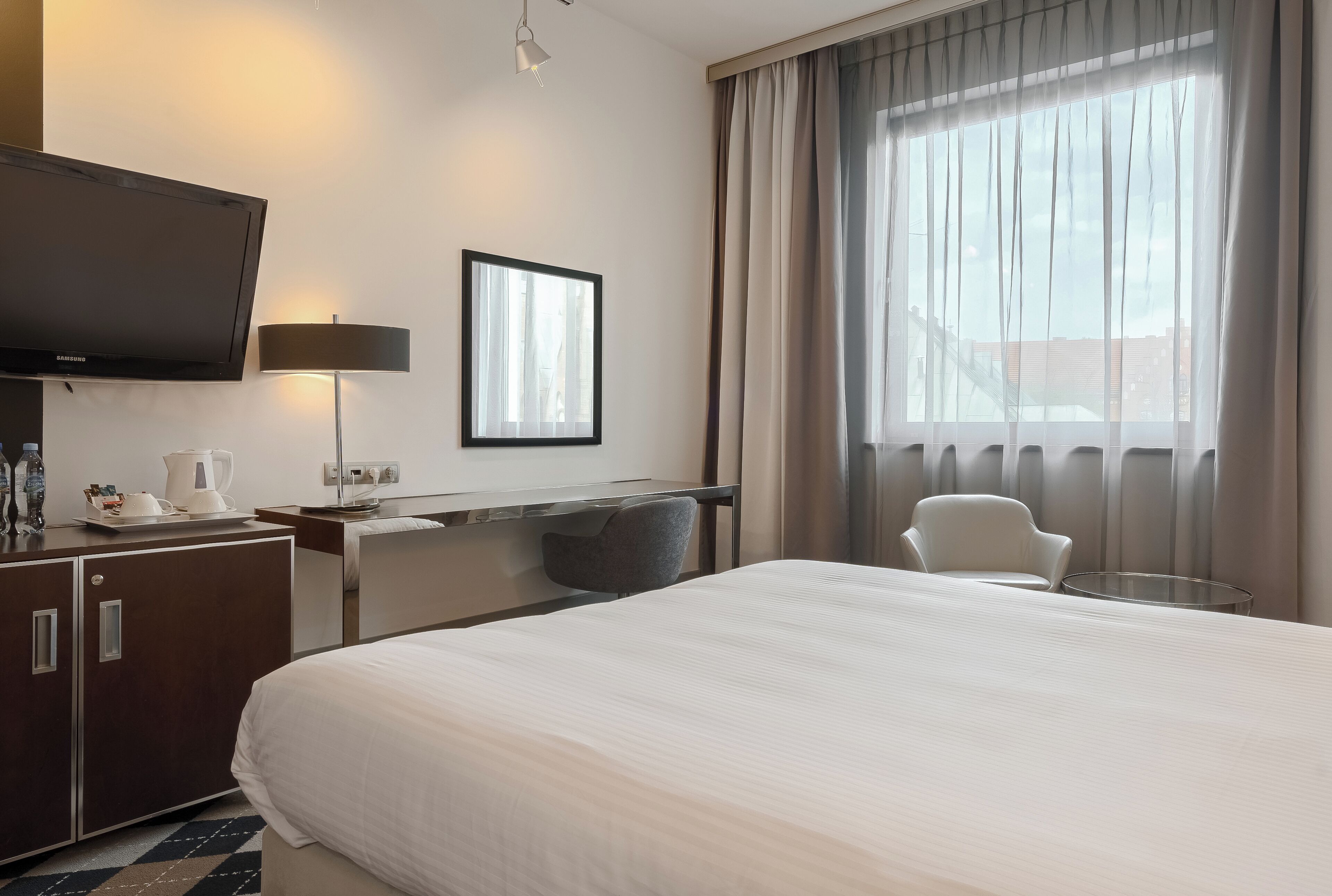 A hotel room with a large bed, a desk with a chair, a TV, a lamp, and a window with curtains.