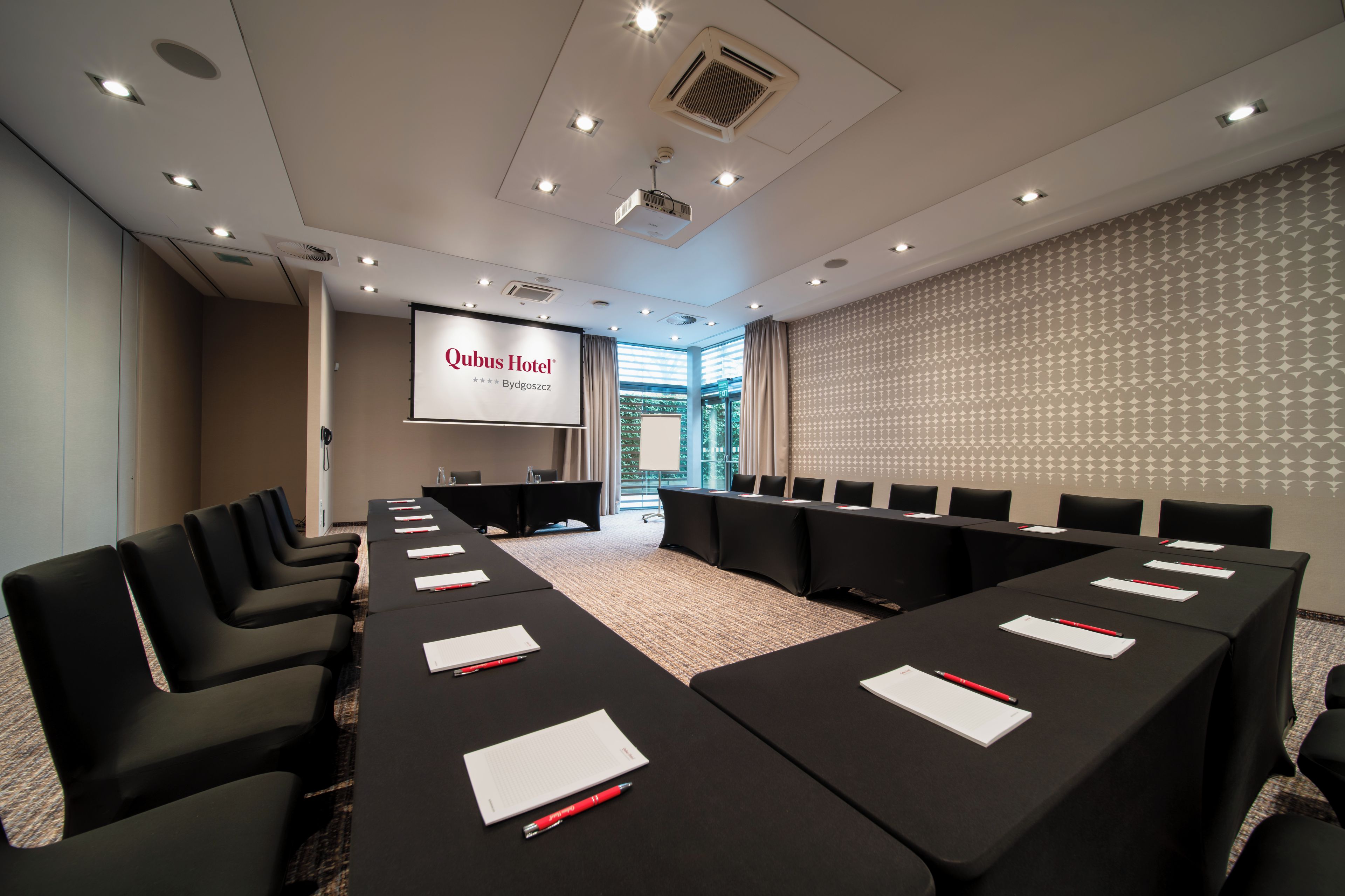 Un salón de conferencias con una larga mesa, sillas, una pantalla de proyector y un papel tapiz estampado.
