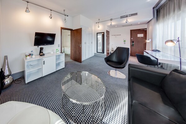 A modern hotel room with a black and white patterned floor, a glass coffee table, a black leather sofa, a white armchair, a TV mounted on the wall, and a desk with a lamp.