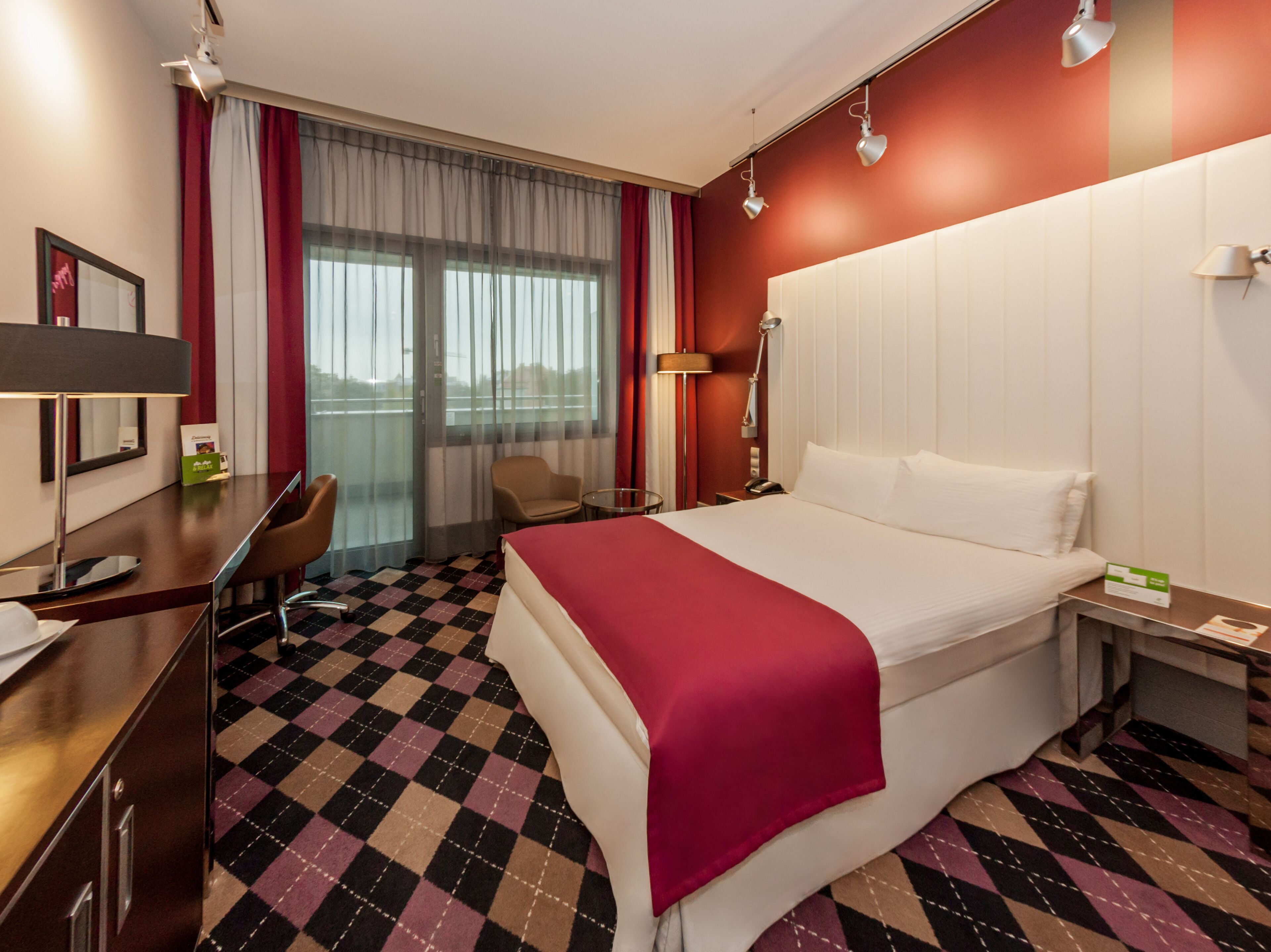 A hotel room with a large bed, a desk with a chair, a small table, and a window with curtains.
