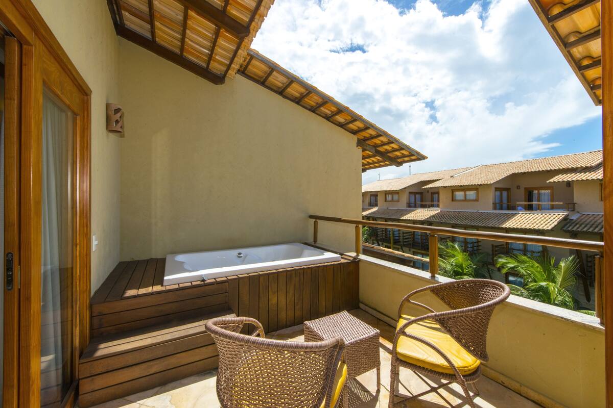 Een balkon met een houten terras, een jacuzzi, rieten stoelen en uitzicht op omliggende gebouwen.
