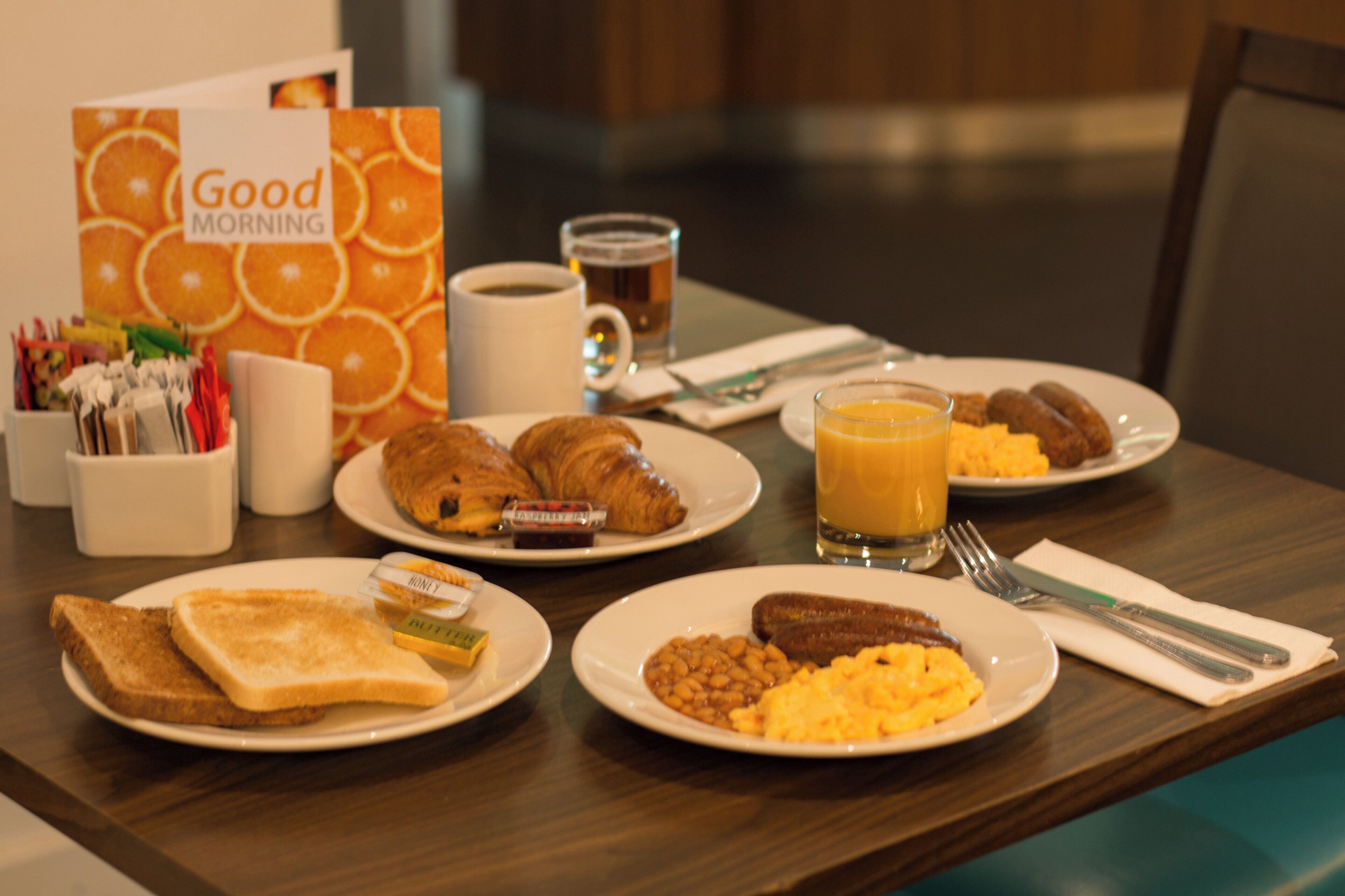 Una mesa de desayuno con tostadas, croissants, huevos revueltos, salchichas y un vaso de jugo de naranja.