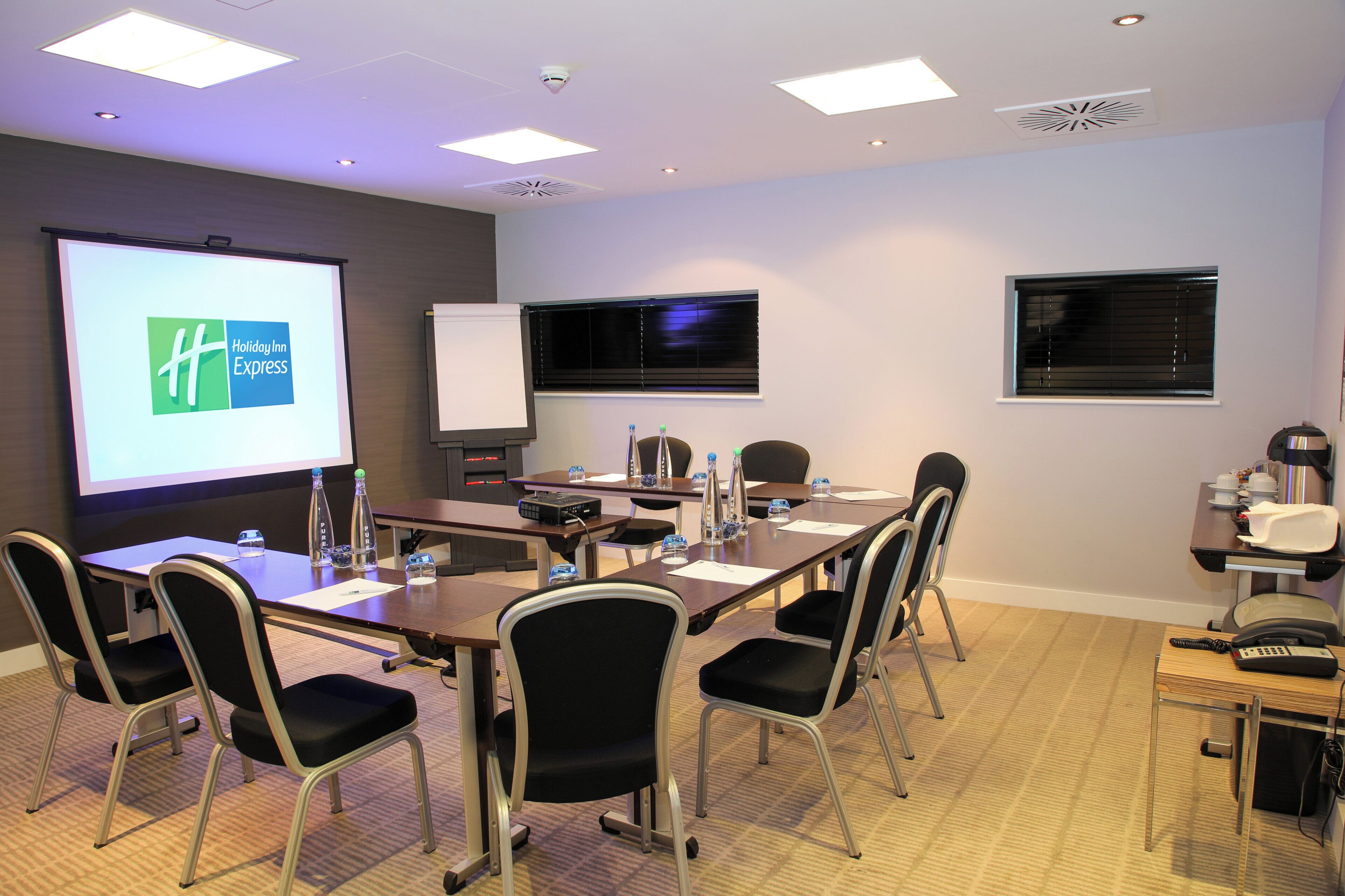 A conference room with a large screen displaying a logo, a projector, a whiteboard, and a table with chairs and water bottles.