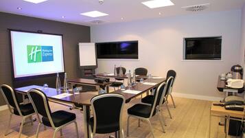 A conference room with a large screen displaying a logo, a projector, a whiteboard, and a table with chairs and water bottles.