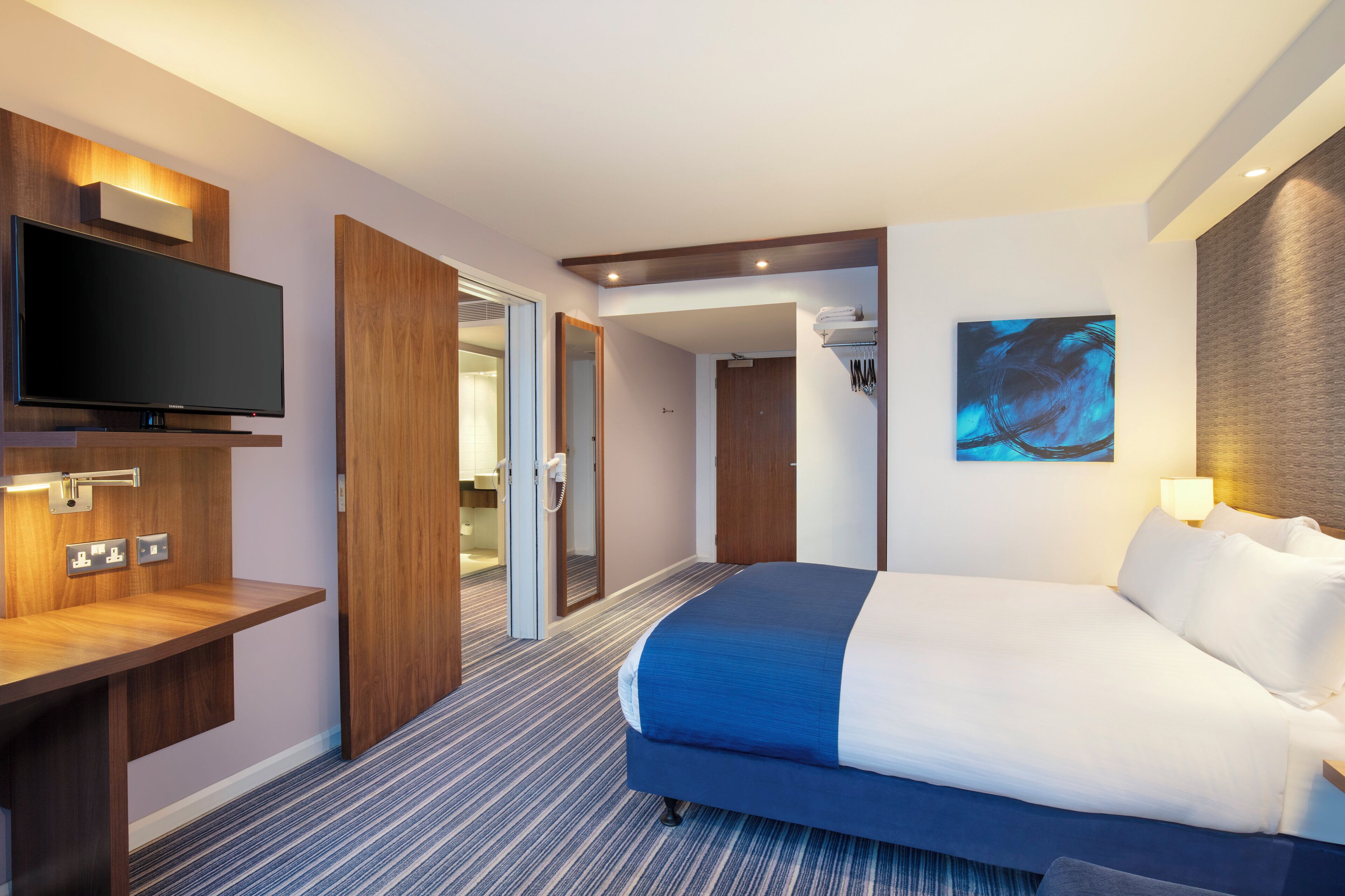 A hotel room with a bed, a television, a desk, and a bathroom visible through an open door.