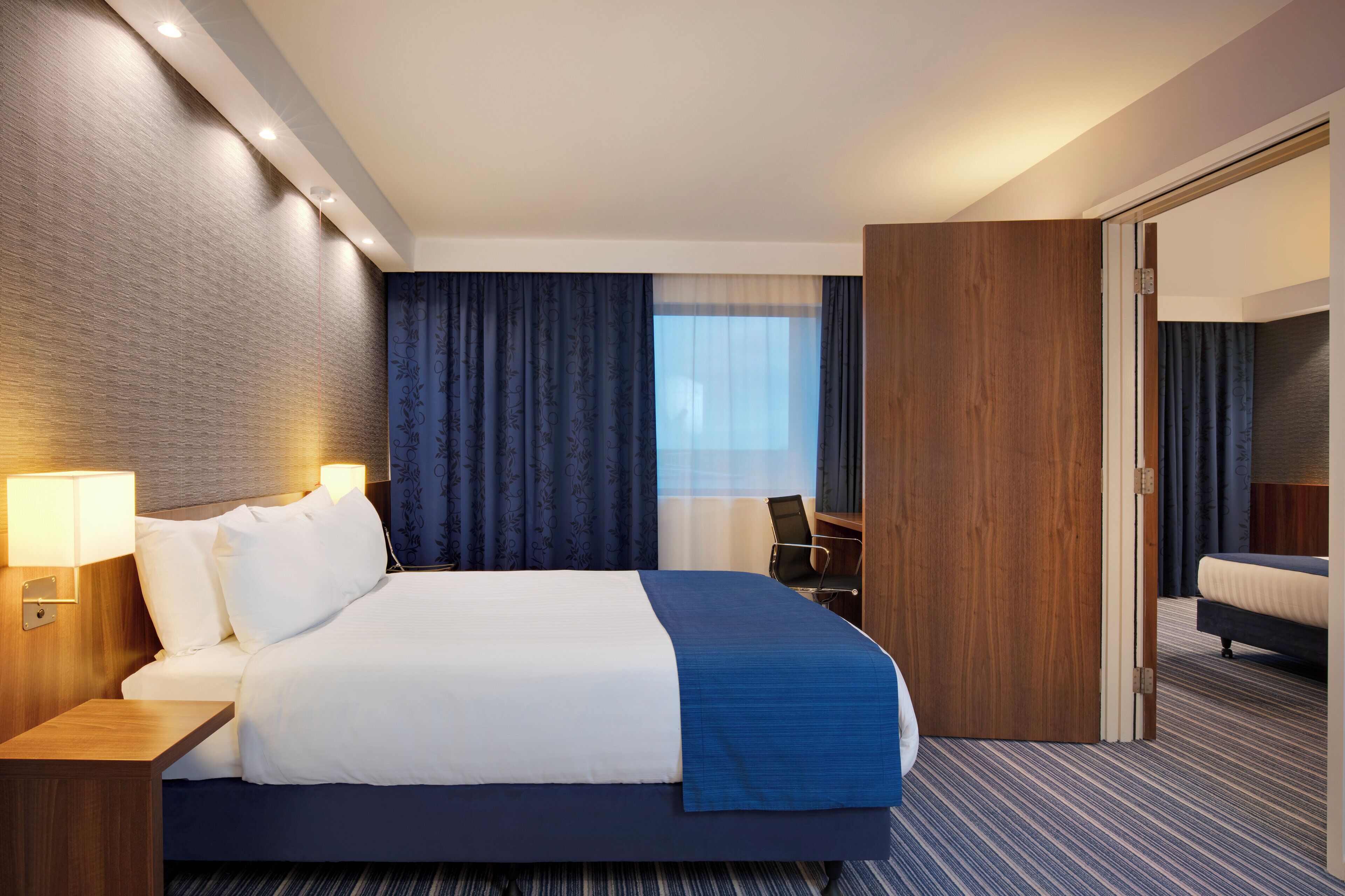A hotel room with a large bed, a desk, a chair, a wardrobe, and a mirror.