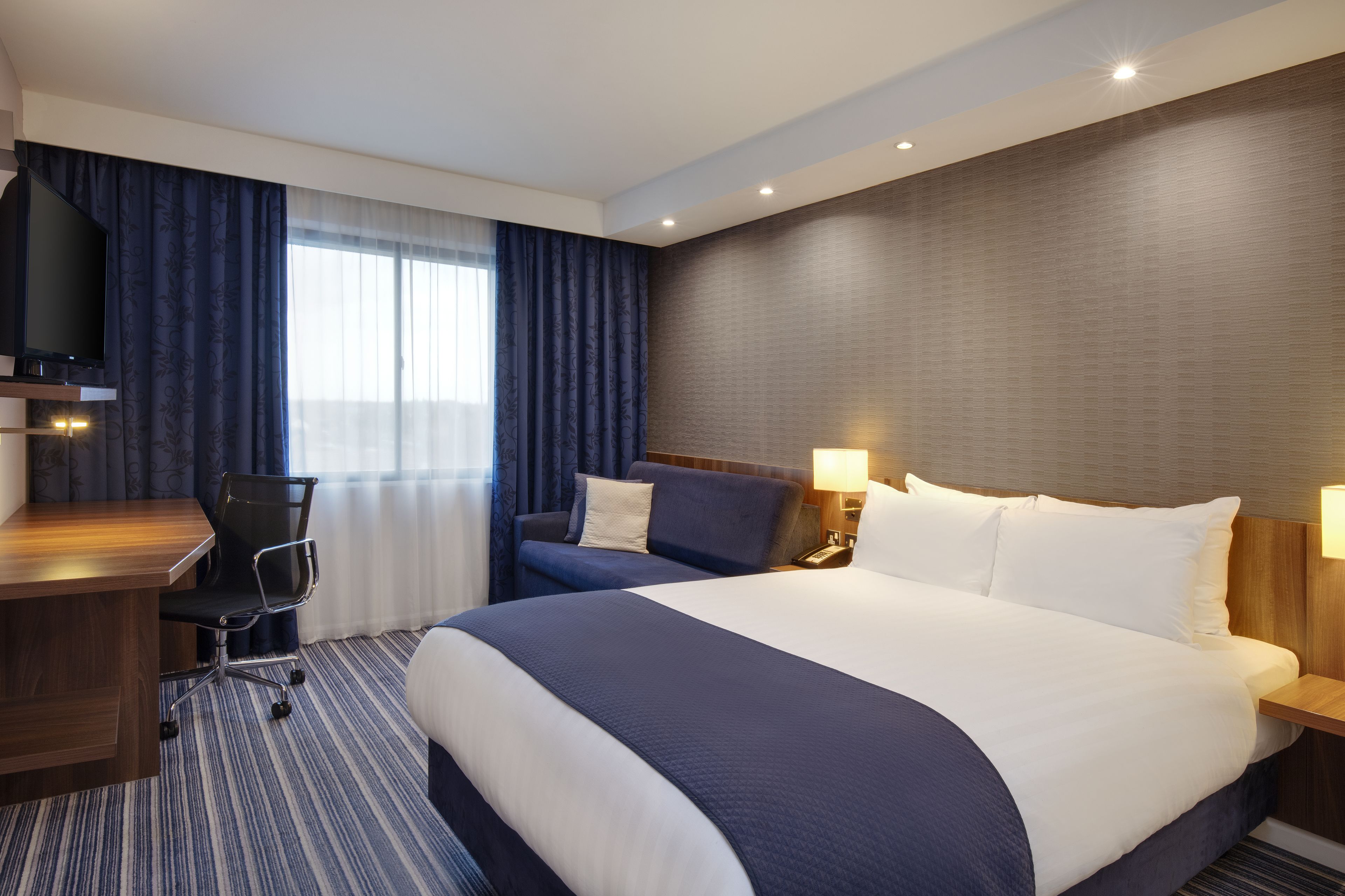 A hotel room with a large bed, a desk, a chair, and a television.