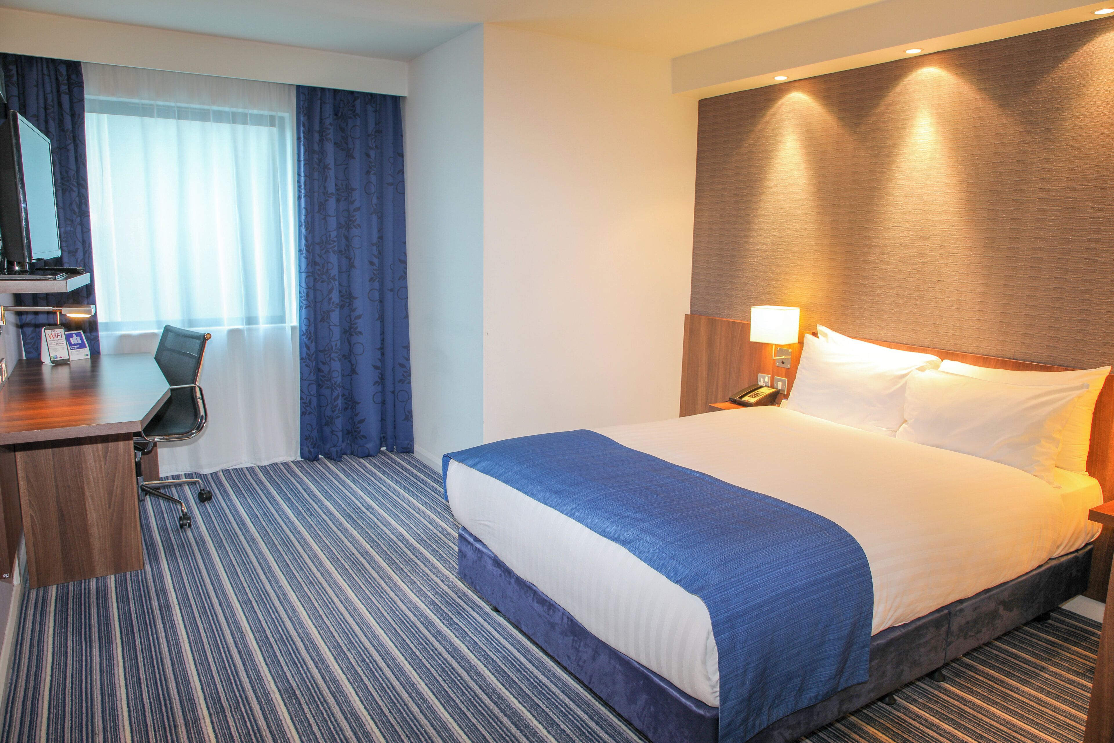 A hotel room with a bed, a desk, a chair, a nightstand, a lamp, and a telephone.